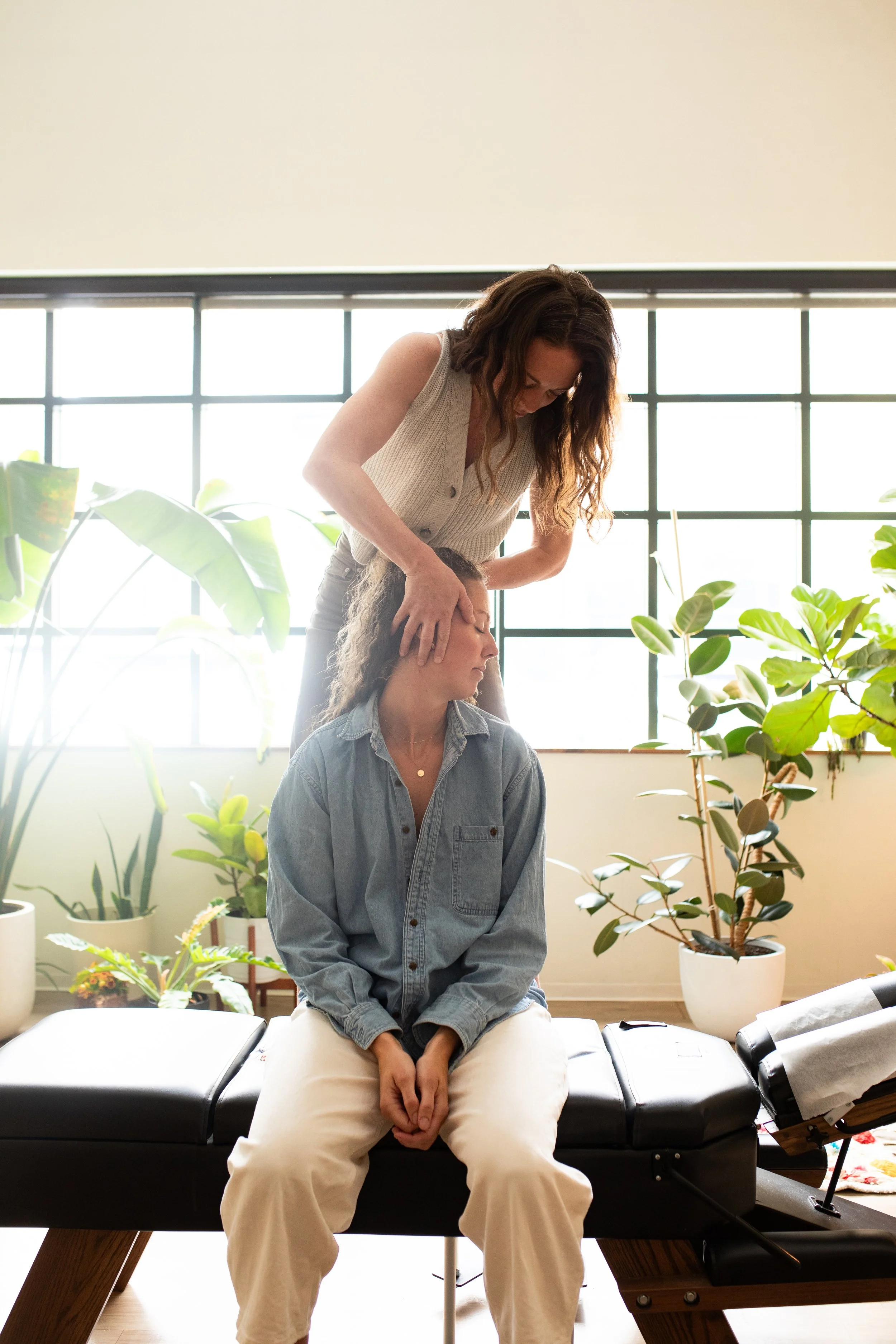 A woman receiving a chiropractic adjustment from a chiropractor in a room with large windows and plants.