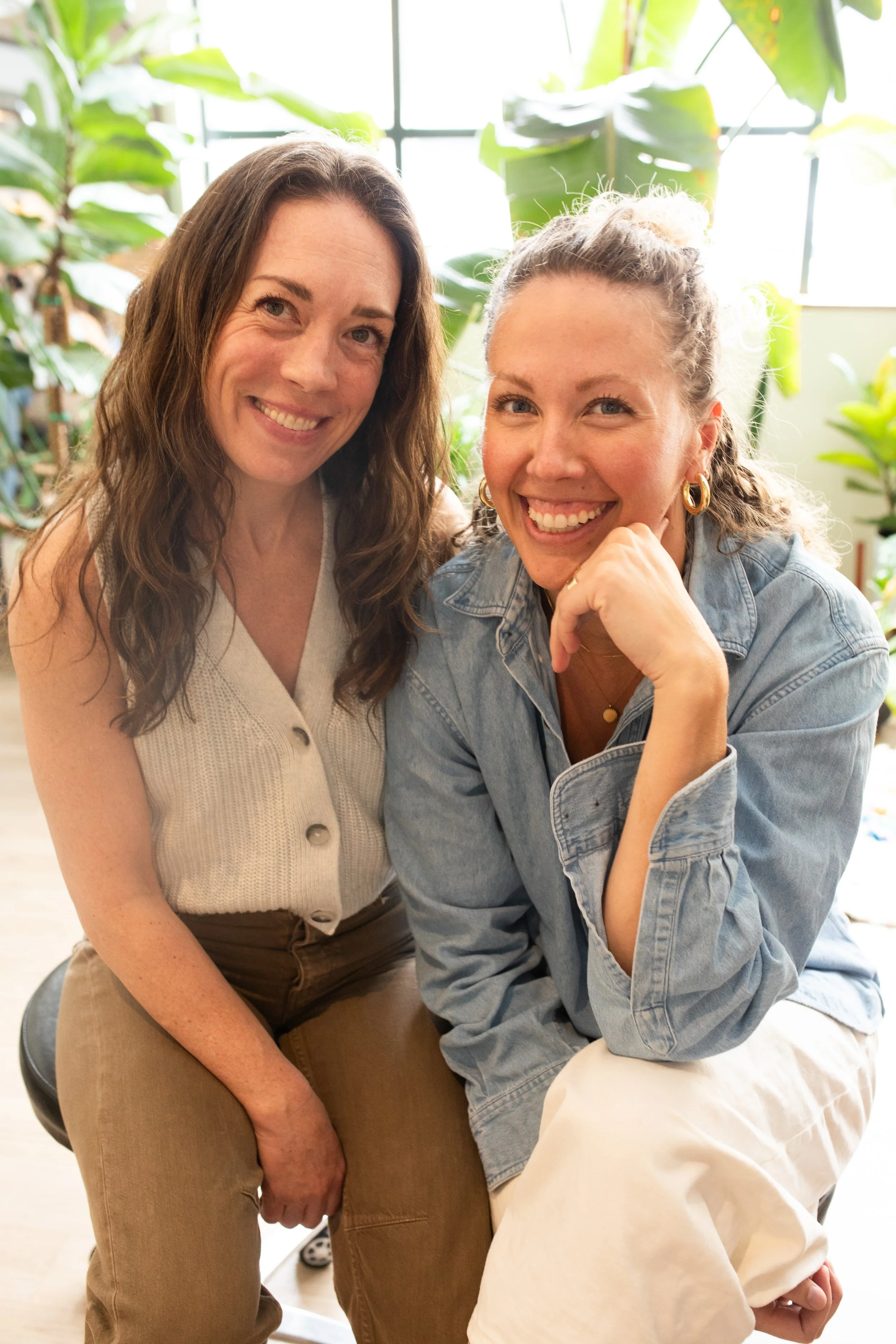 Two women smiling and sitting close together indoors, surrounded by green plants and natural light.