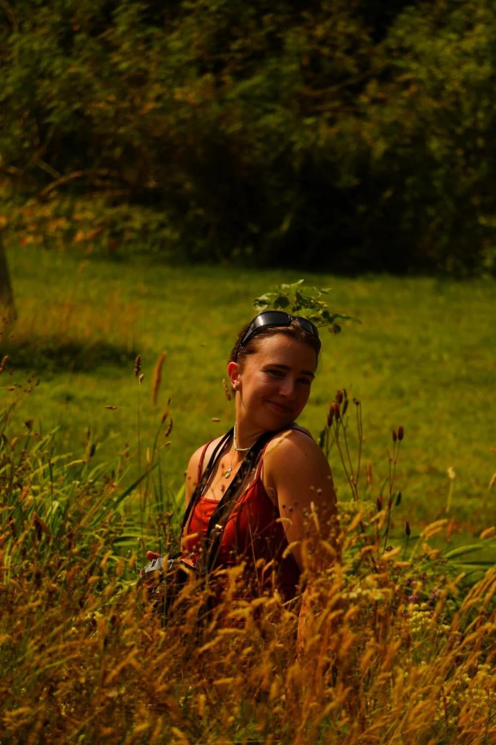 A woman standing in a field of tall grass and wildflowers, with trees in the background, smiling and looking over her shoulder.