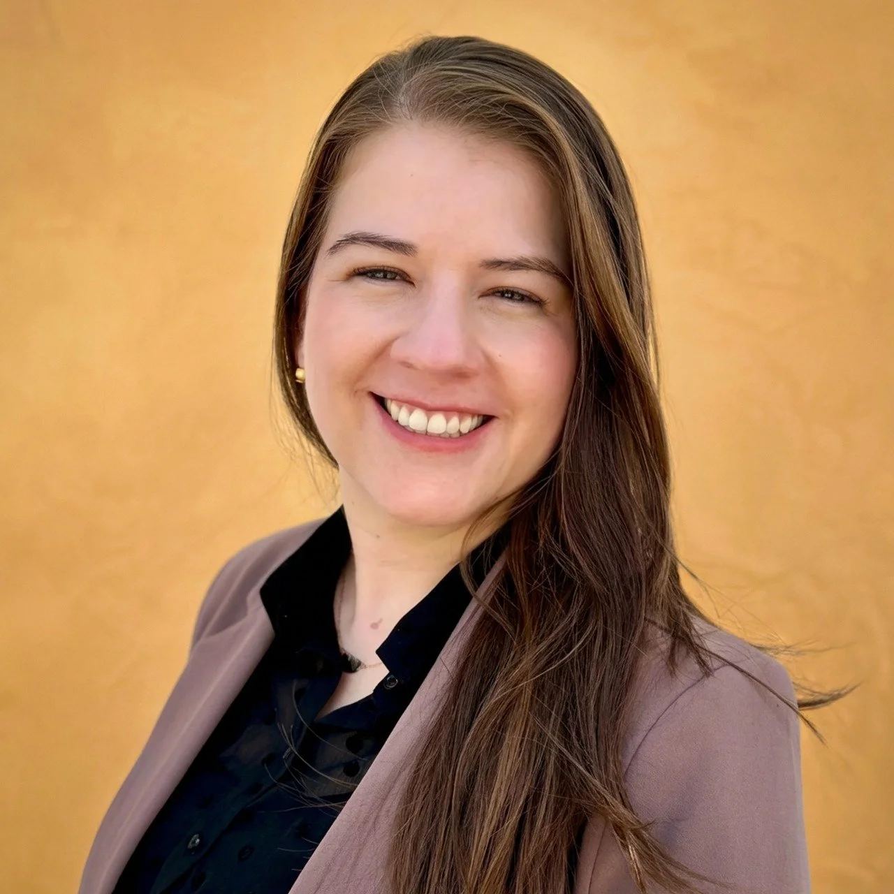 An image of Yenny Paez, smiling,  wearing a black top and a light brown blazer, with a yellow-colored background.