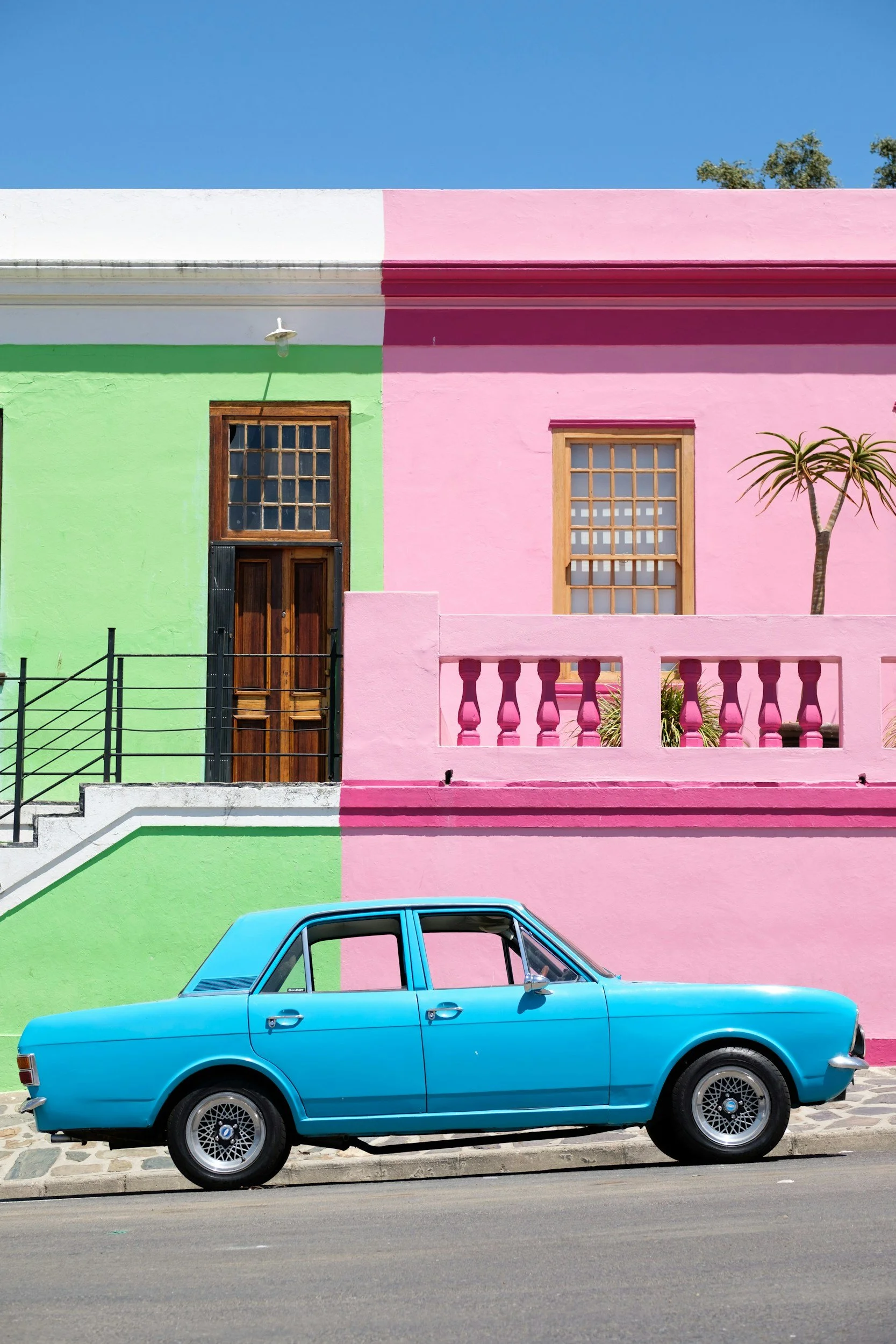 A blue vintage car parked in front of a building with a green and pink facade under a clear blue sky.