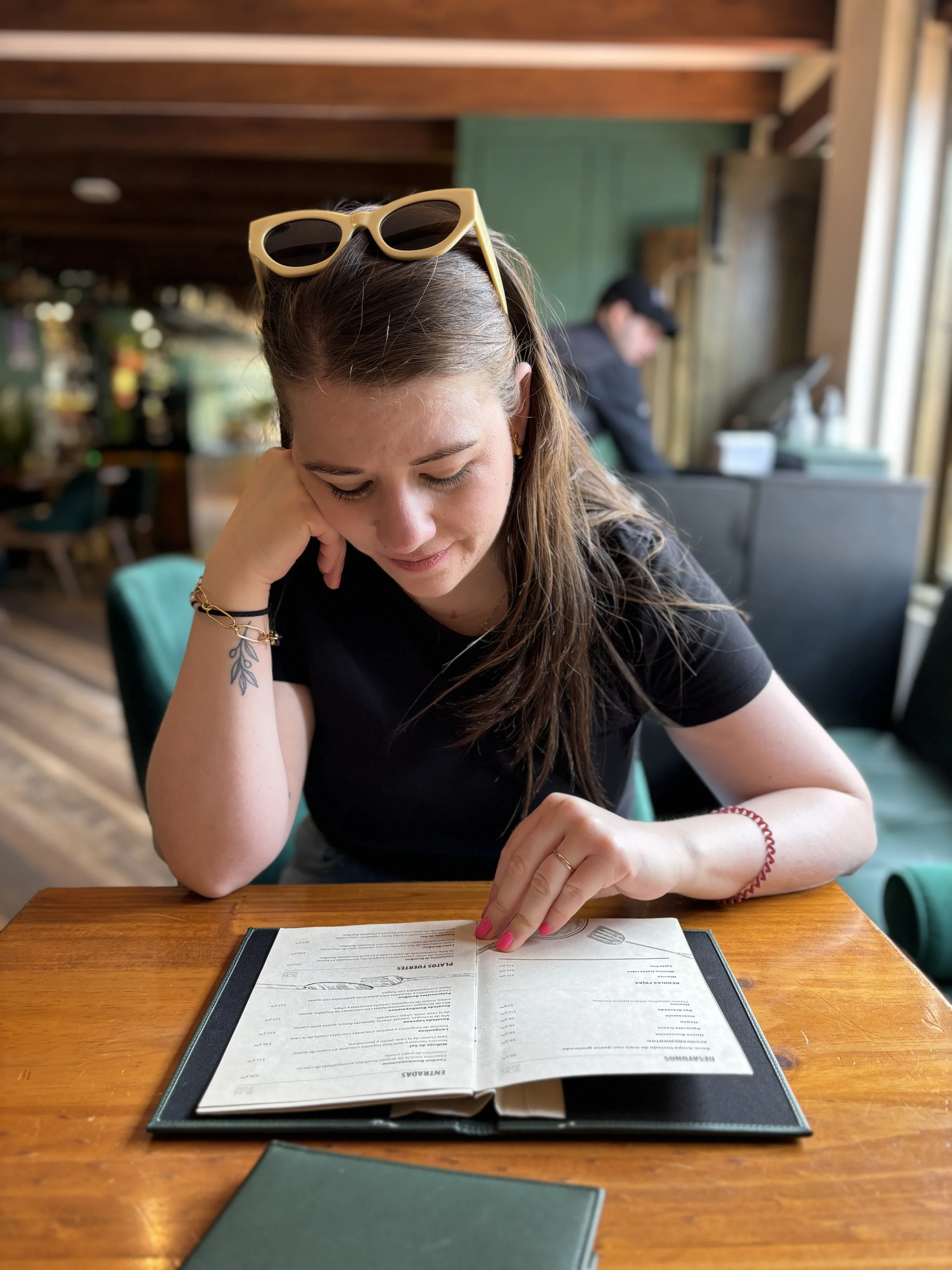 Yenny wearing sunglasses on her head, a black t-shirt, and jewelry, sitting at a wooden table in a restaurant and looking at a menu.