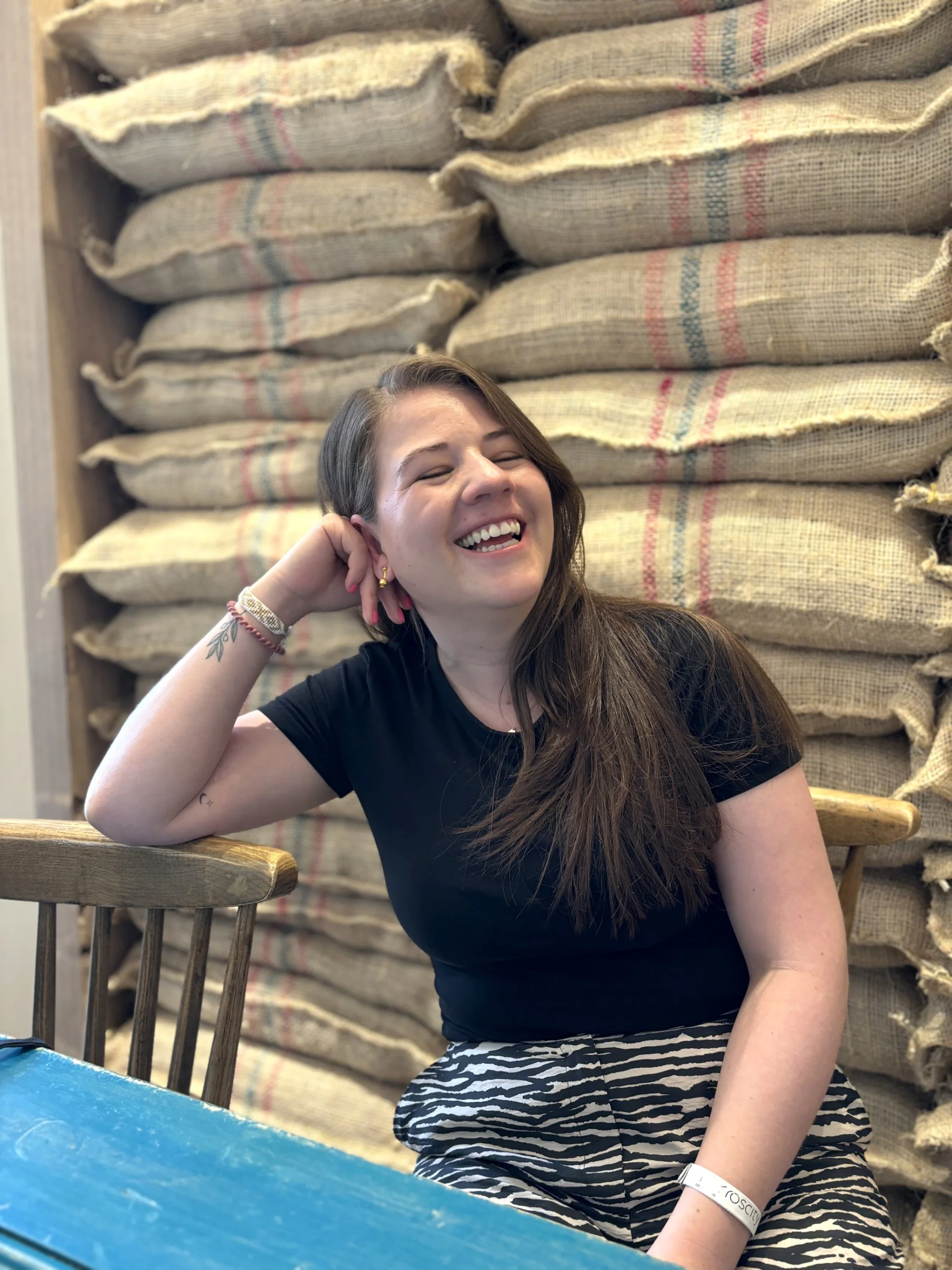 A young woman laughing and smiling with her eyes closed, sitting at a wooden table against a background of stacked burlap sacks.