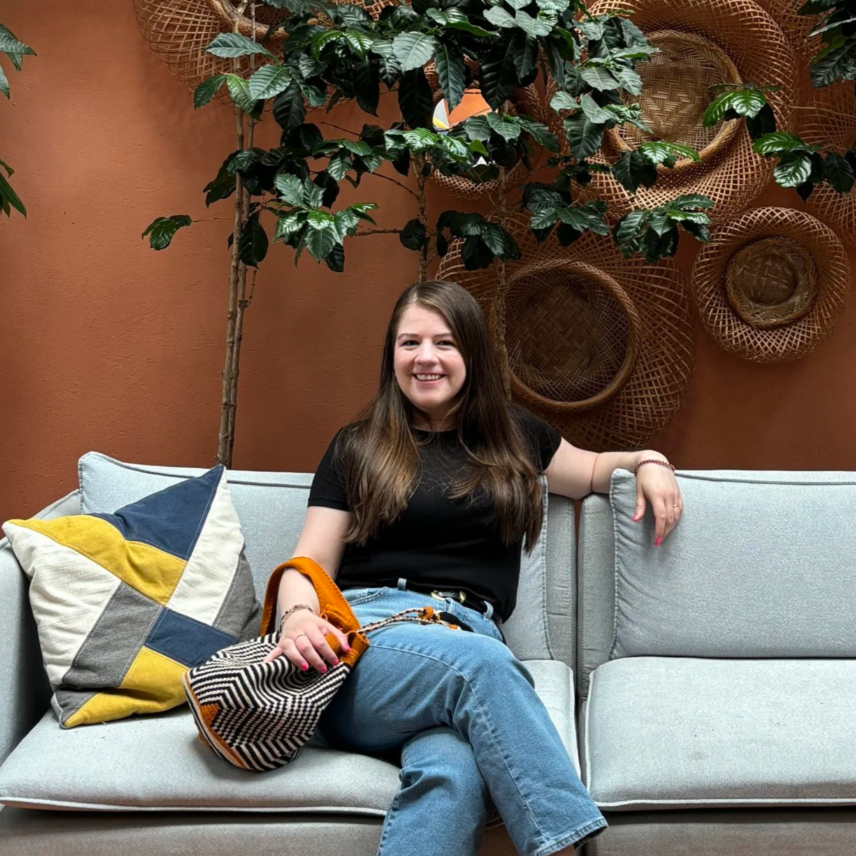 A woman with long brown hair sitting on a light gray sofa, smiling at the camera. She is wearing a black t-shirt and blue jeans, with a multicolored handbag resting on her lap. Behind her is a rust-colored wall adorned with woven baskets and greenery.