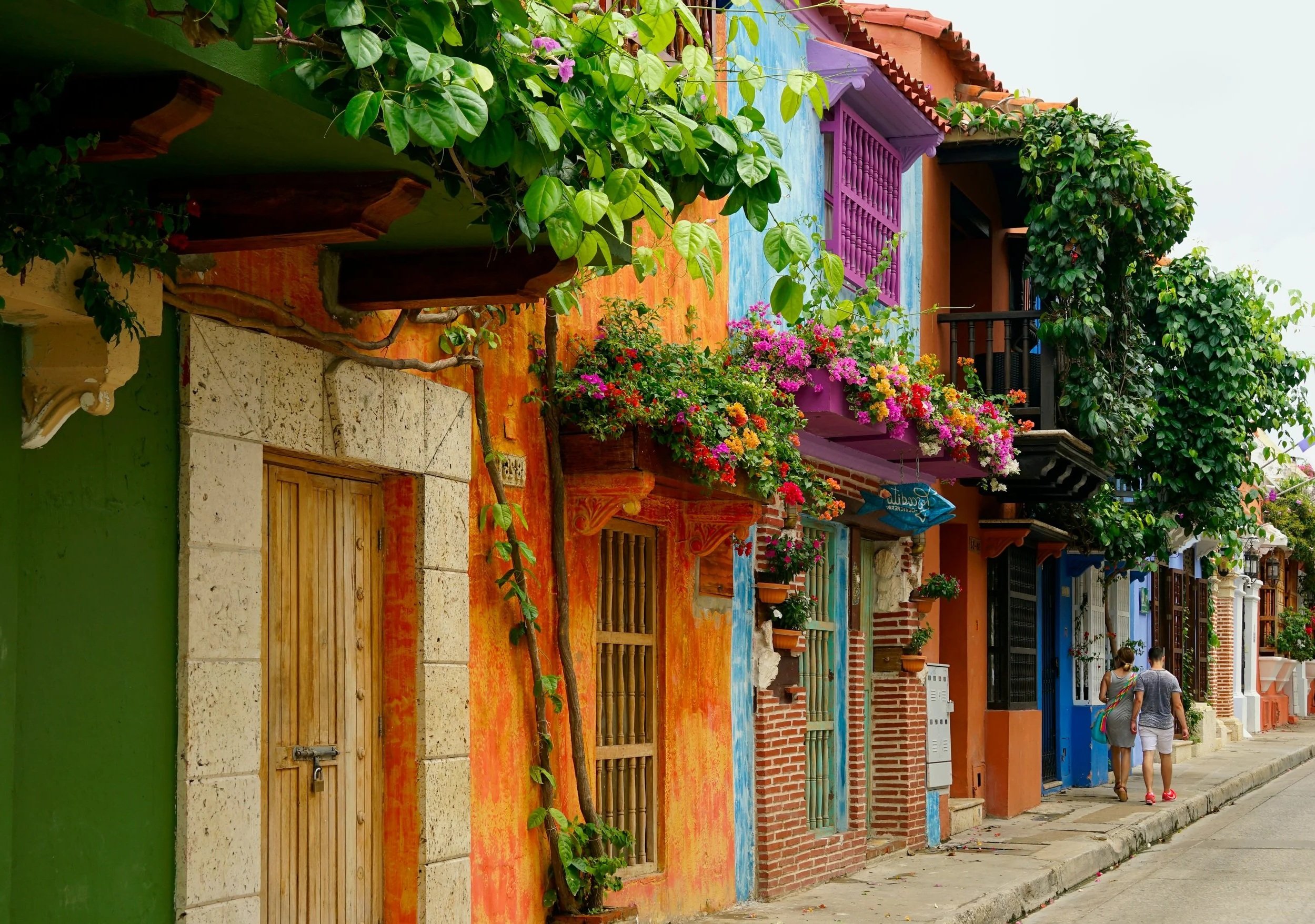 Colorful street with vibrant buildings decorated with flowers and greenery, two people walking on the sidewalk.