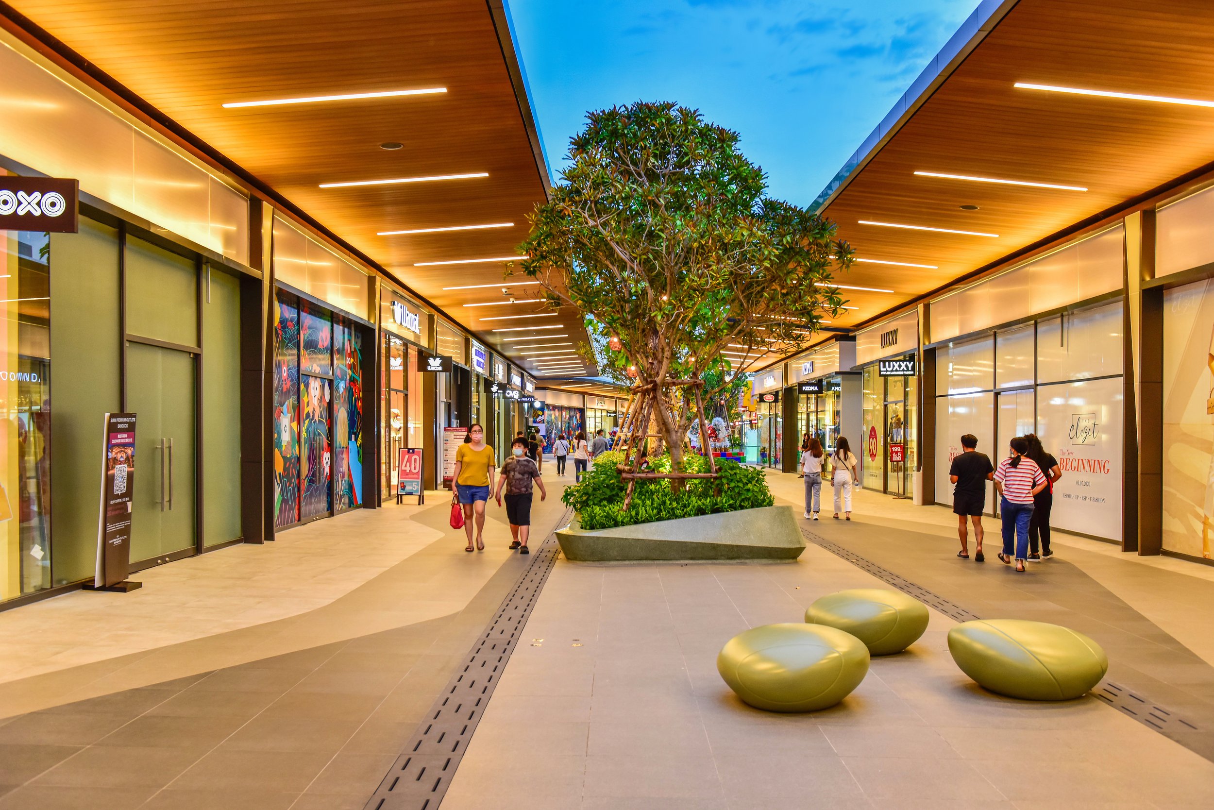Interior of a shopping mall with a centerpiece tree, shoppers walking, and store storefronts on both sides.