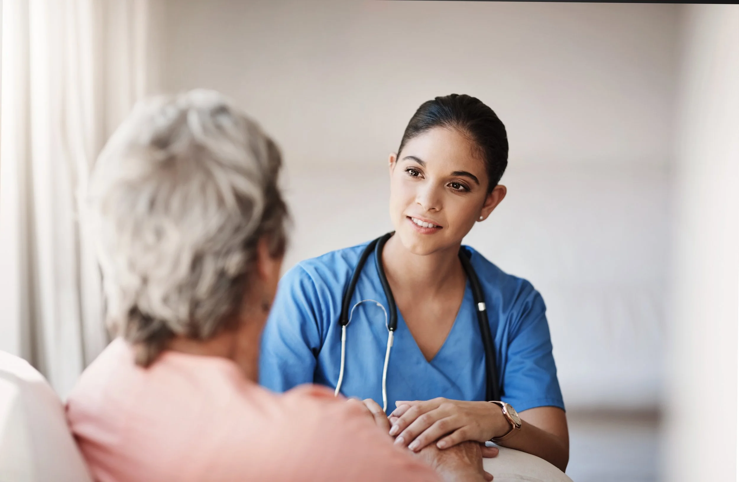 A female healthcare professional in blue scrubs with a stethoscope around her neck talking to an older patient in a hospital bed.