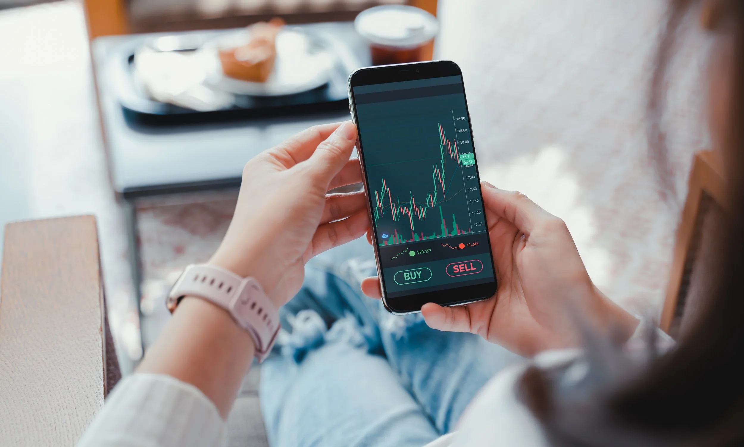 Person holding smartphone displaying stock trading app with buy and sell buttons, showing a candlestick chart and market data.