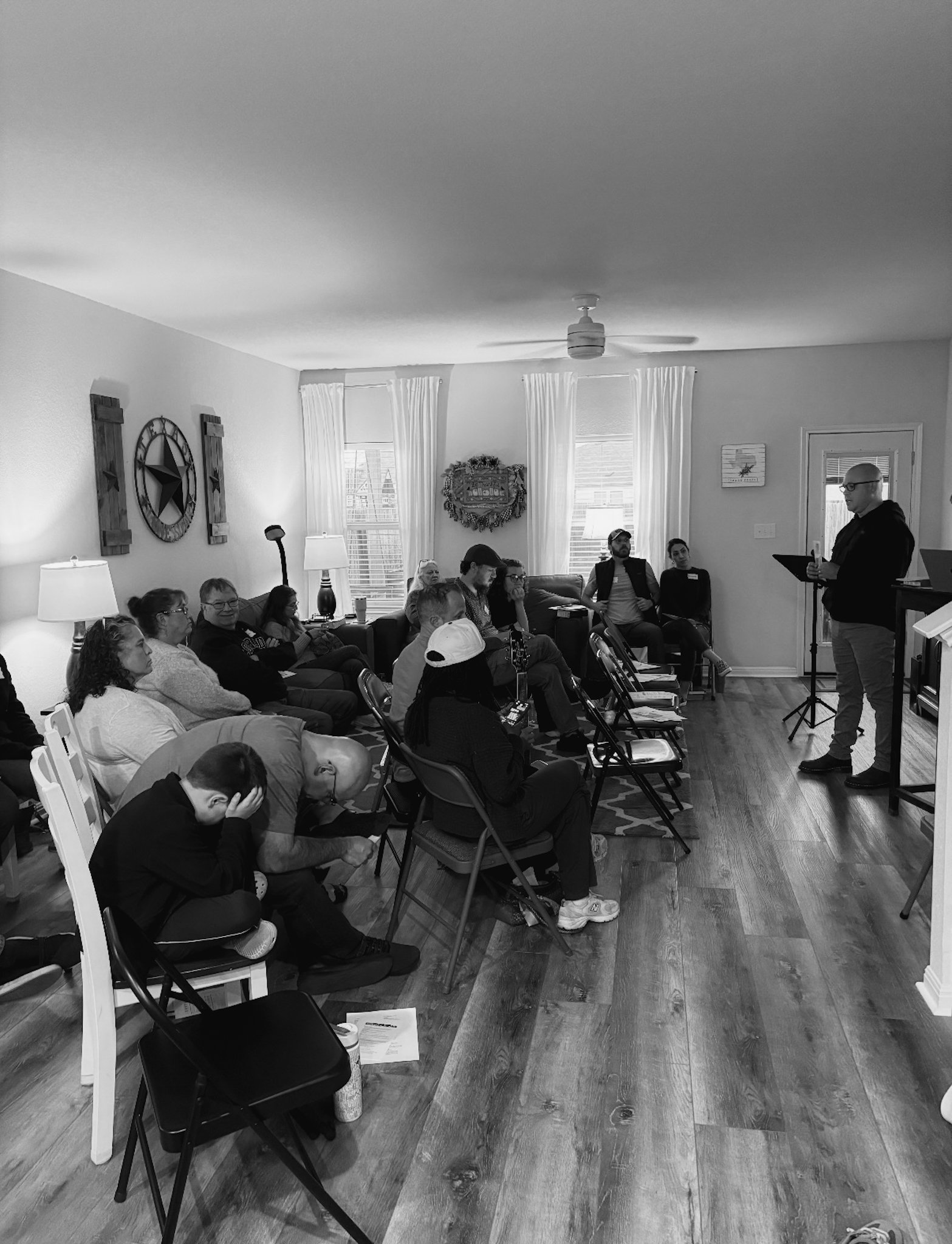 A group of people seated in a living room, listening to a person standing and speaking at a podium, with some taking notes and others looking on.