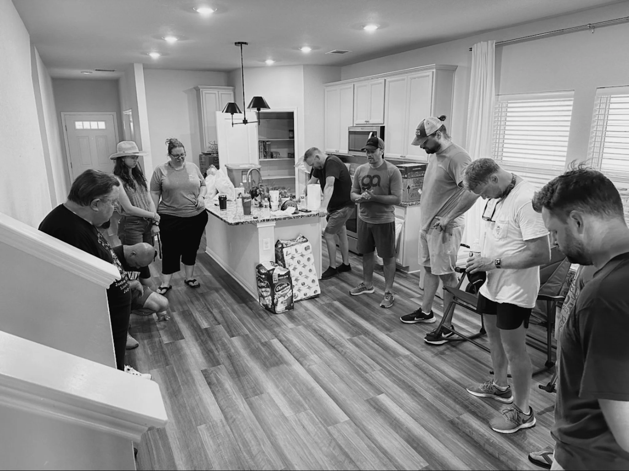 A group of people in a kitchen appear to be praying, with heads bowed and eyes closed, before a meal or gathering.