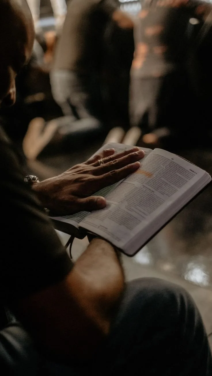 A person sitting in a dark room reading a large open book, possibly in a church or auditorium during a service or event.