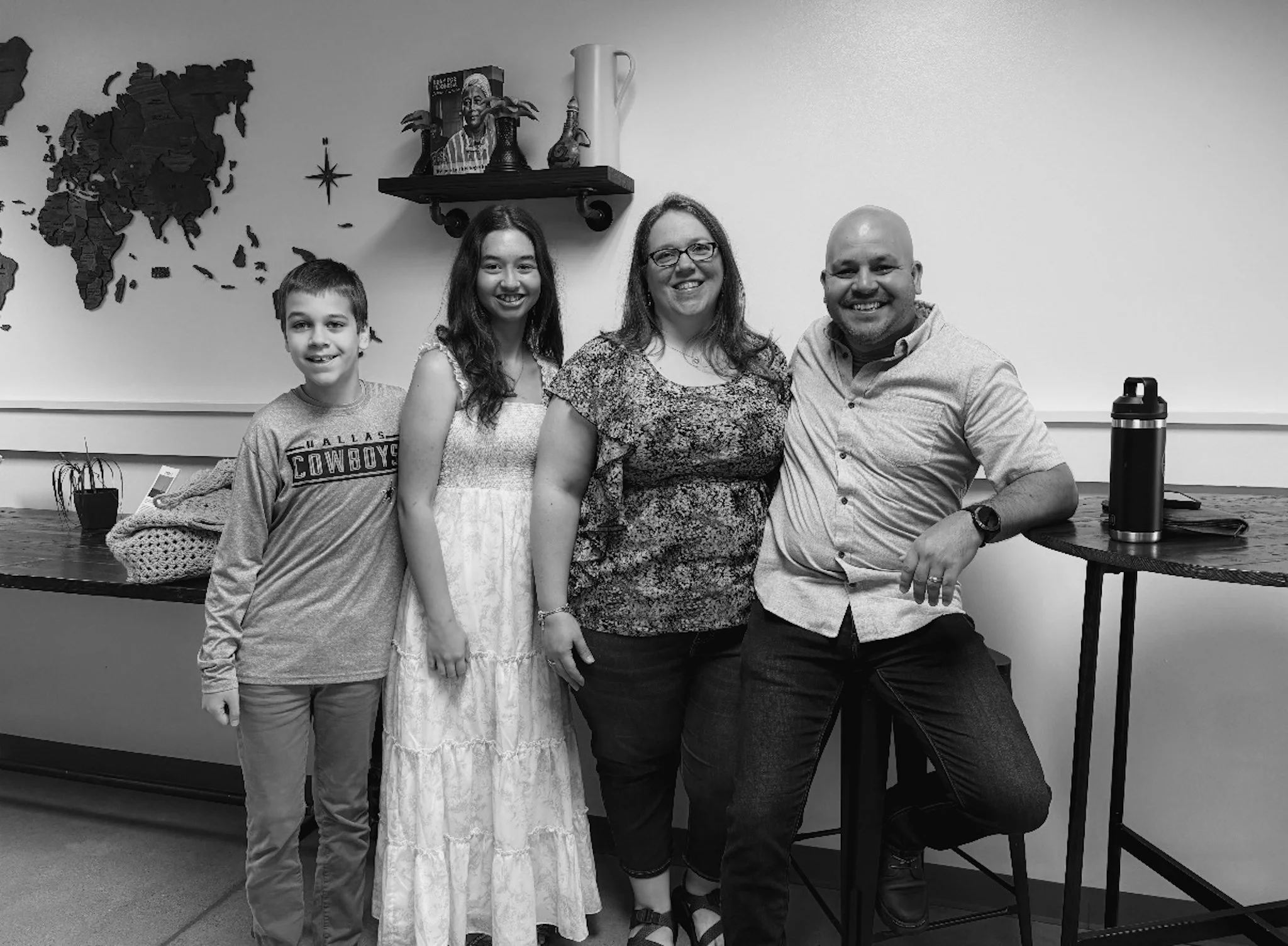 A family of four standing indoors, smiling at the camera. The background includes a world map wall art, a shelf with decorations, and a table with a water bottle.