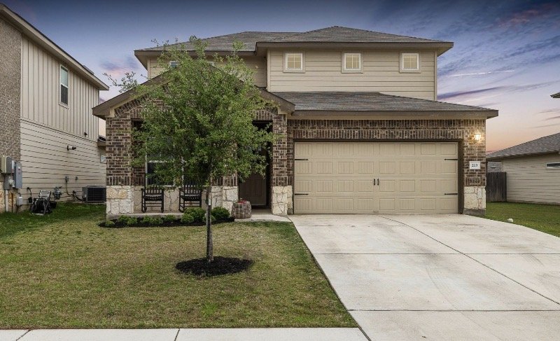 Two-story house with beige siding and brick accents, a lawn with a small tree, a concrete driveway, and a two-car garage, during sunset.