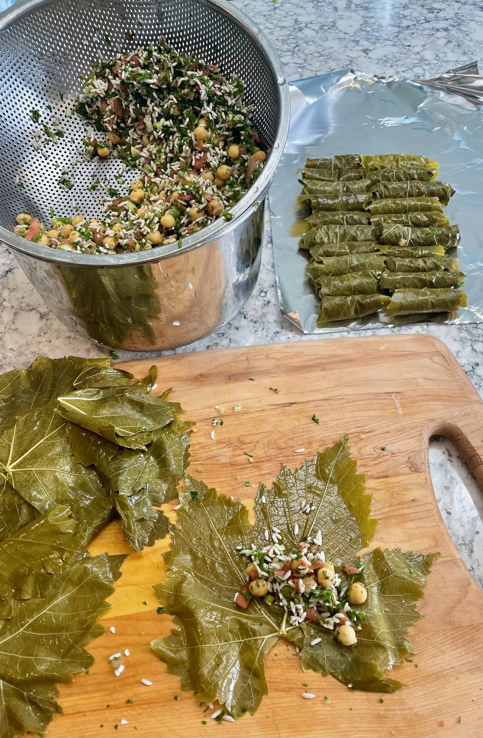A kitchen countertop with a metal bowl of chickpea salad, a cutting board with grape leaves filled with rice, and loose grape leaves.