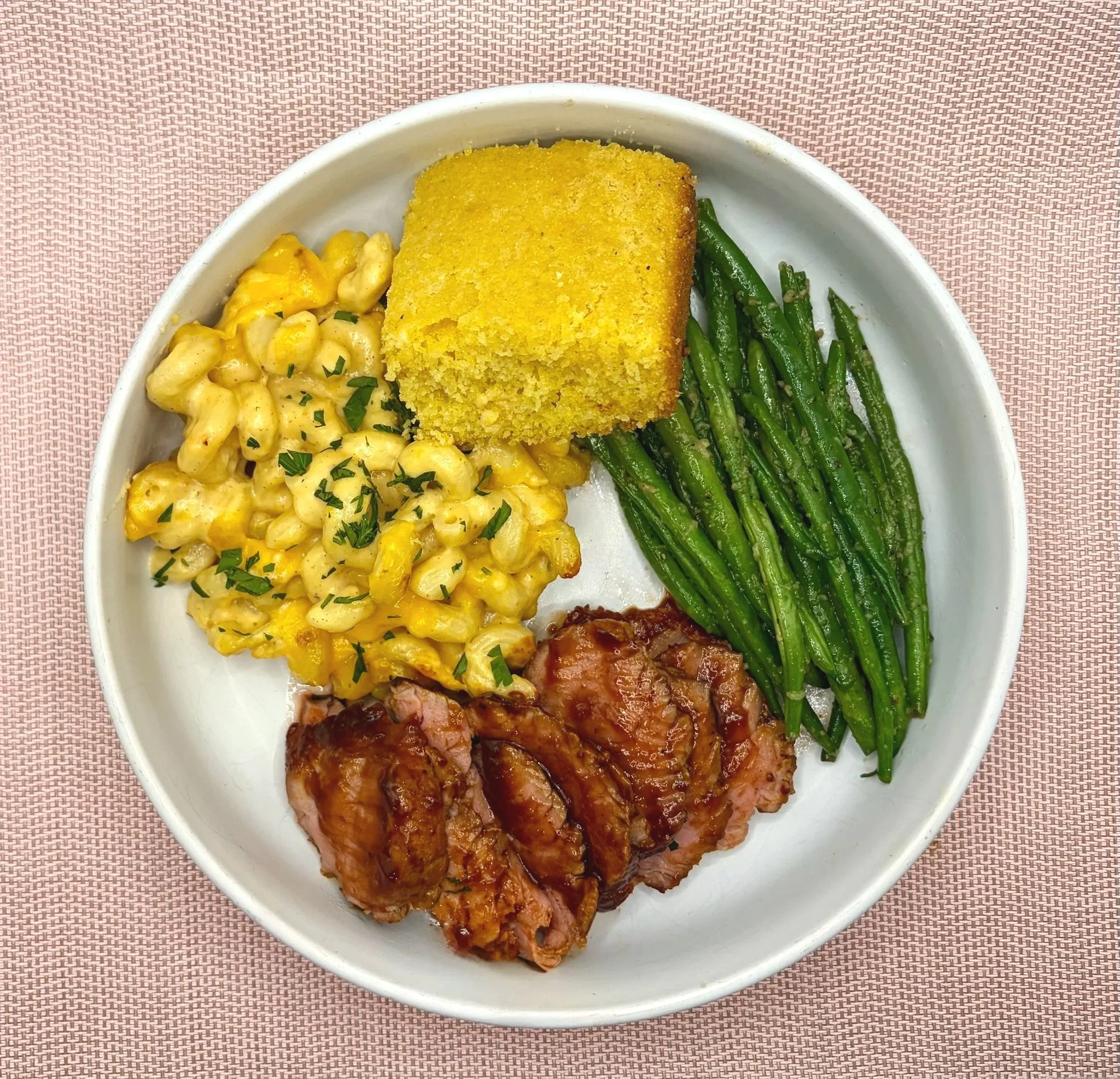 A white bowl containing golden cornbread, macaroni and cheese topped with chopped parsley, green beans, and pieces of glazed ham, placed on a pink woven placemat.