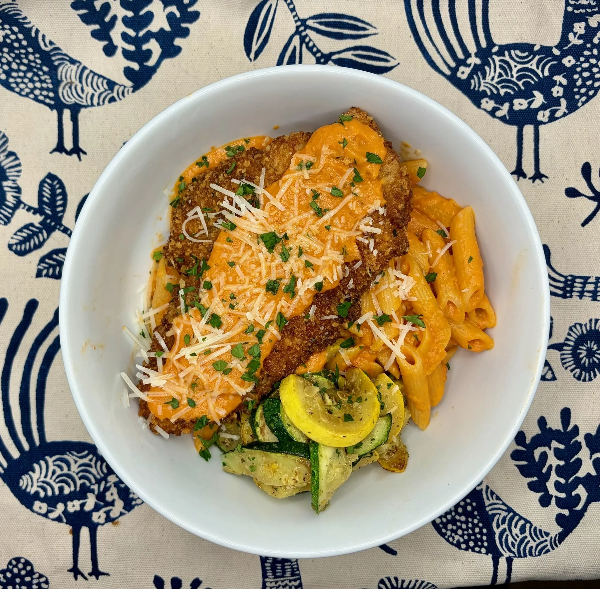Bowl of macaroni and cheese topped with shredded cheese, chopped parsley, and a breaded chicken cutlet on a blue and white patterned tablecloth.