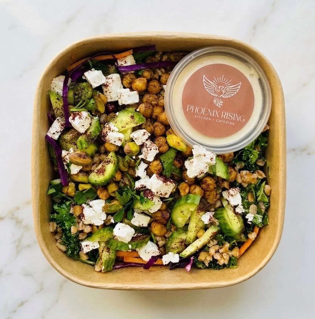 A salad in a brown cardboard takeout bowl with mixed greens, sliced avocado, chickpeas, purple cabbage, crumbled feta cheese, and a small container of dressing from Phoenix Rising Kitchen & Catering.