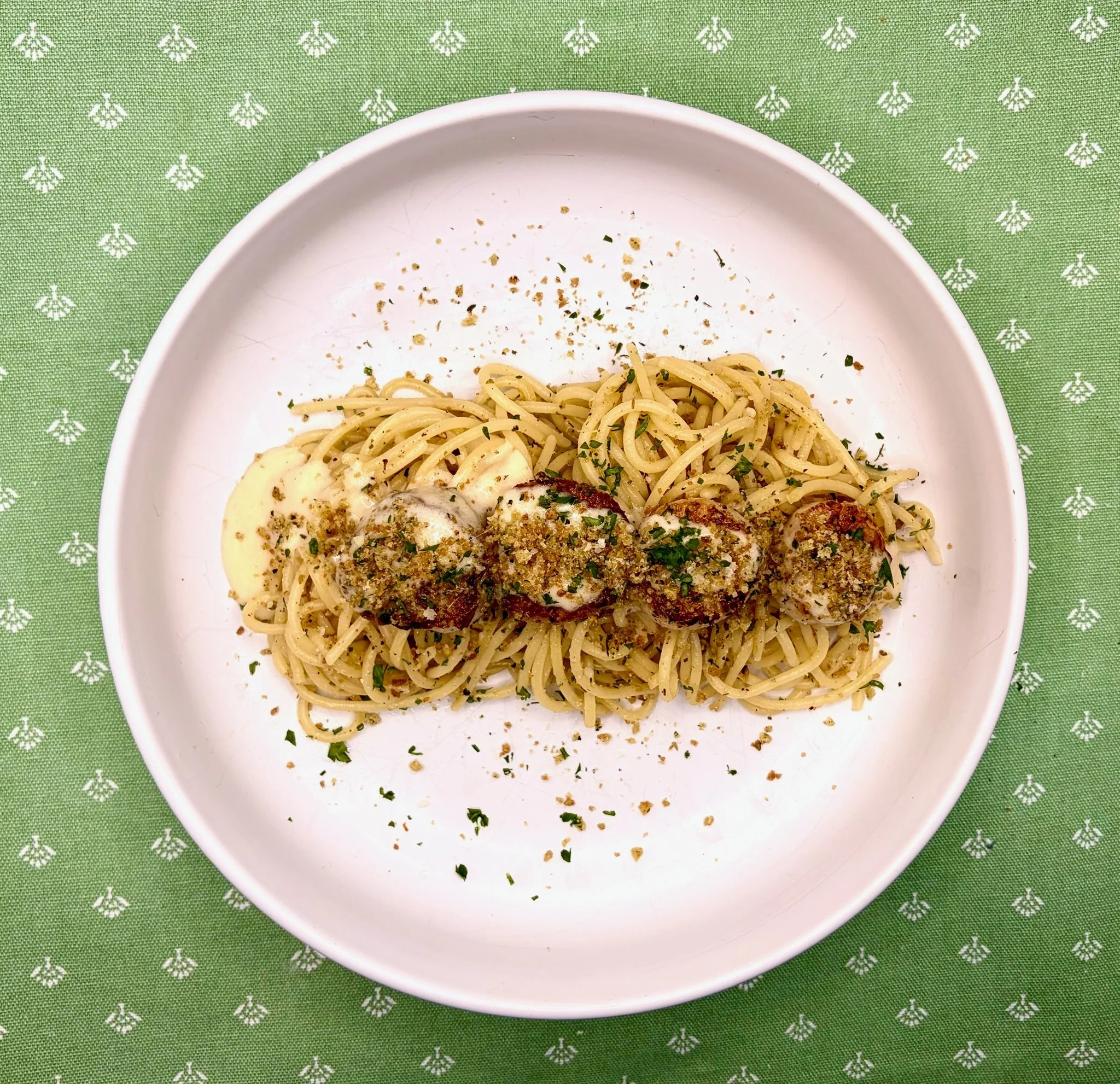 A white bowl of spaghetti garnished with chopped herbs and crushed red pepper, placed on a green tablecloth with a white floral pattern.