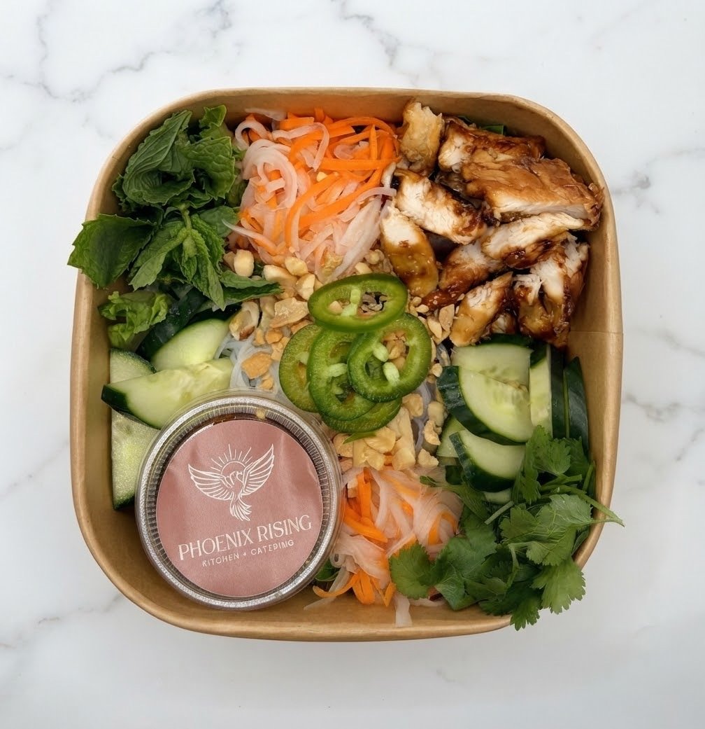 Healthy salad bowl with grilled chicken, cucumber, jalapenos, shredded carrots, shredded white radish, lettuce, cilantro, crushed nuts, and a small container of dressing, all inside a brown takeout container on a white marble surface.