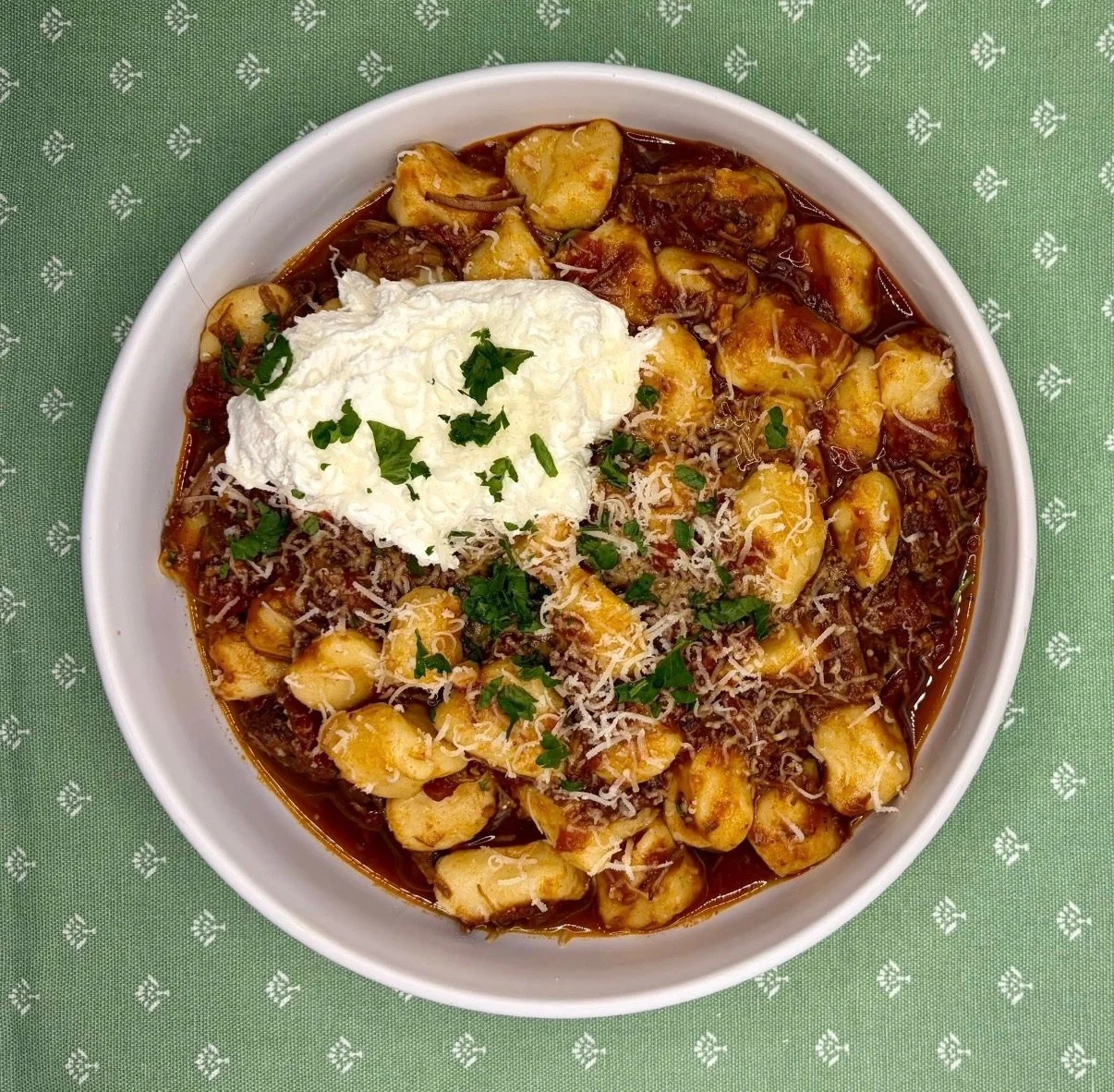Bowl of chicken and dumplings with tomato-based broth, garnished with sour cream, shredded cheese, chopped green herbs, served on a green tablecloth.