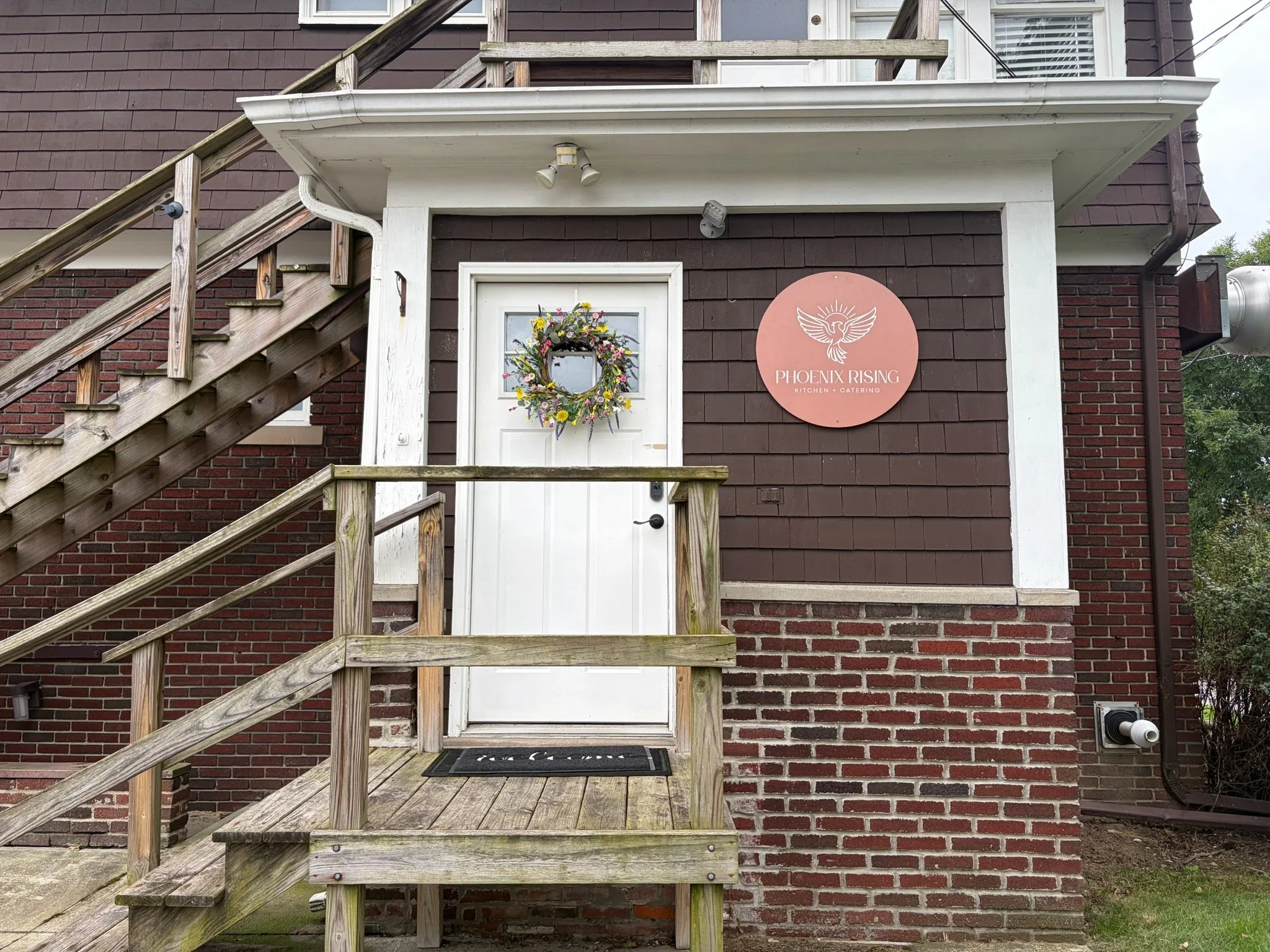 The image shows the location of Phoenix Rising Kitchen. There is an outdoor staircase with wooden railings on the left side leading to an upper level. The building has brown and red brick walls with white trim.