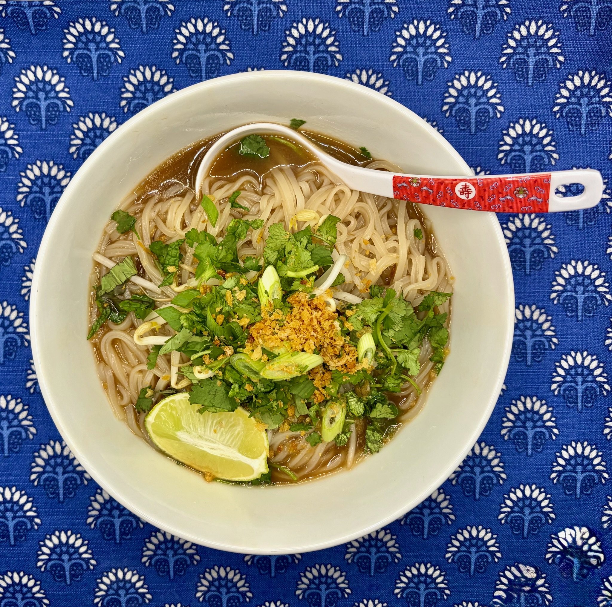 A bowl of noodle soup with cilantro, green onions, and a lime wedge, placed on a blue patterned tablecloth.
