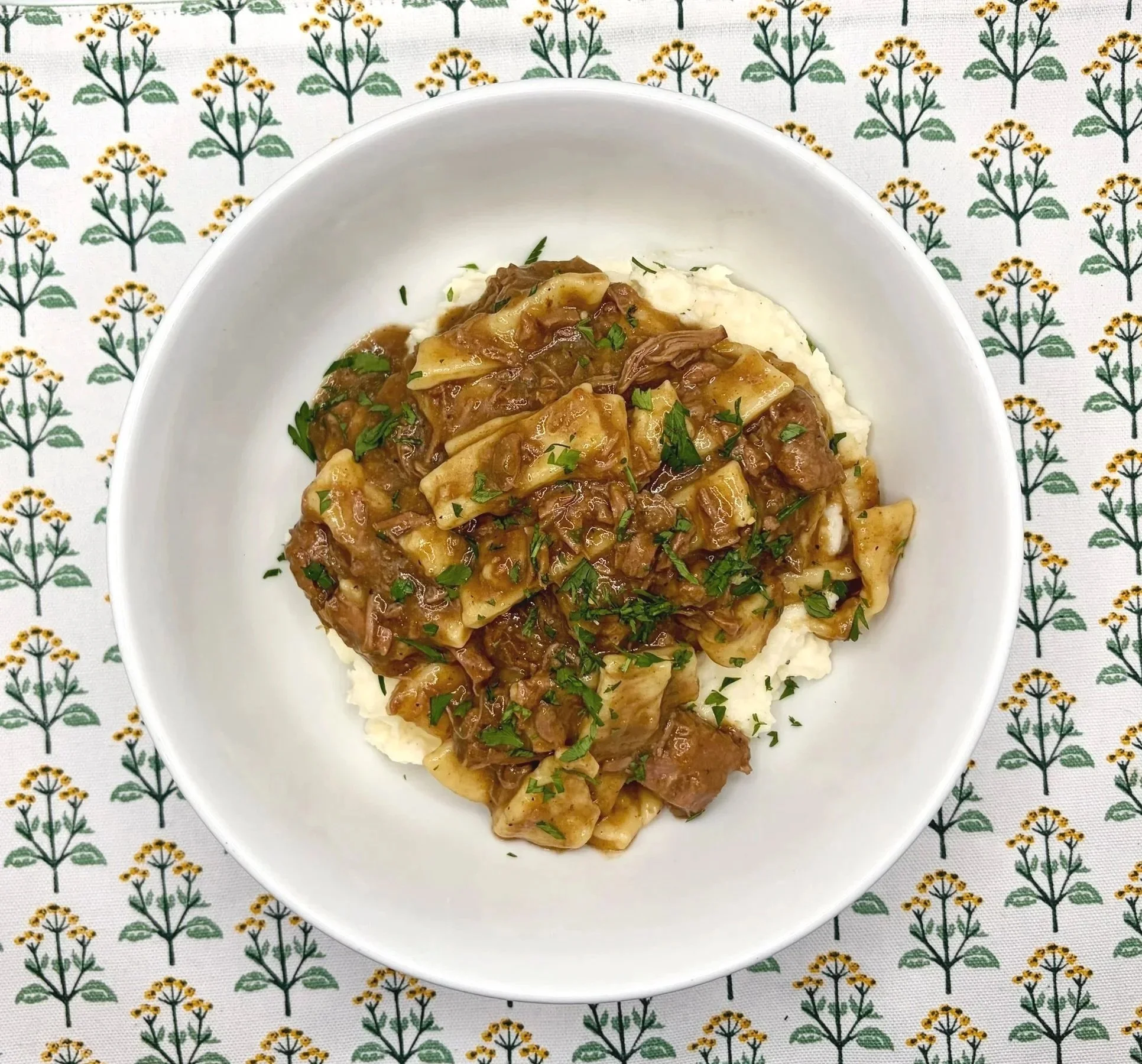A white bowl of beef stew with mashed potatoes on a floral tablecloth.