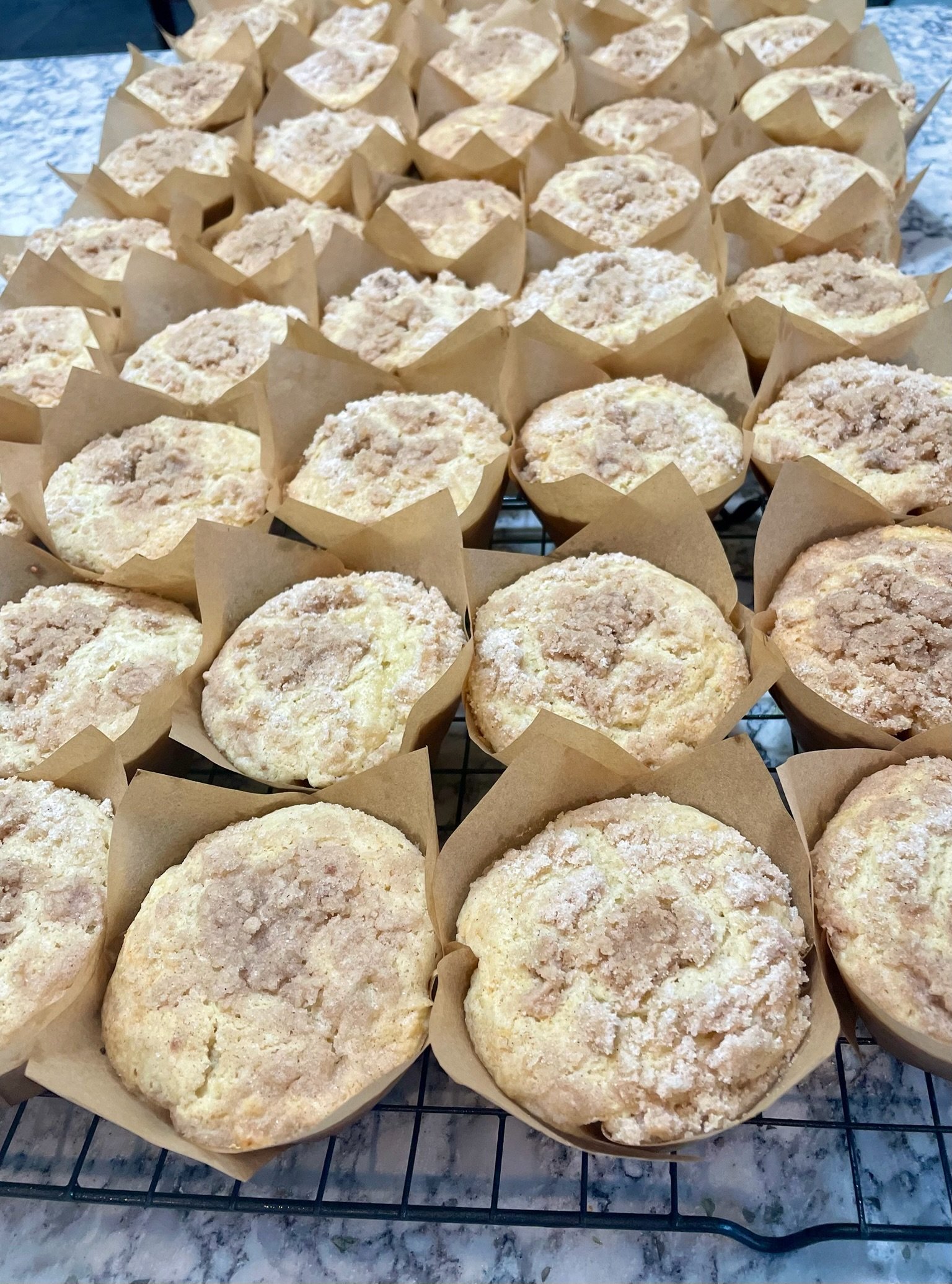 Multiple muffin-like baked goods on a cooling rack, each in a paper cup, with a crumbly topping, on a marble countertop.