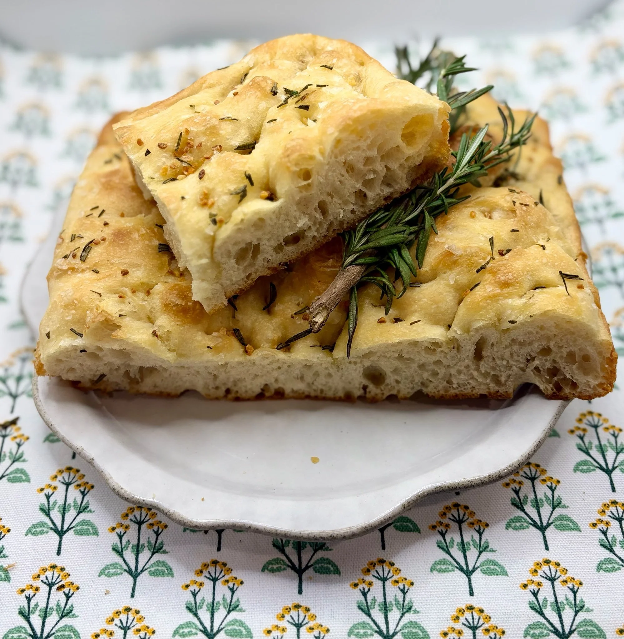 Close-up view of freshly baked focaccia bread seasoned with rosemary and herbs on a decorative tablecloth.