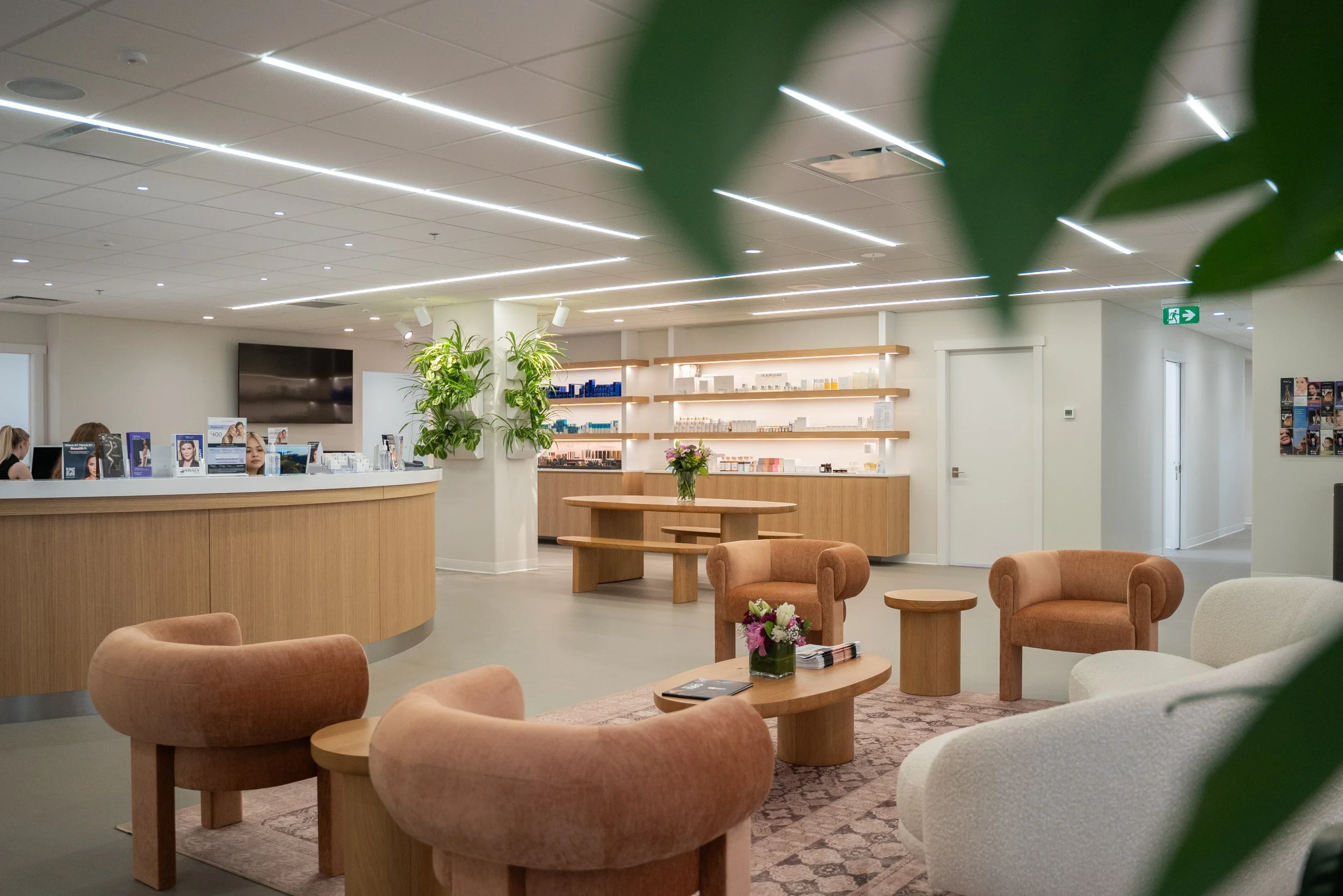 A modern waiting area or lobby with beige and white furniture, wooden accents, potted plants, and shelves with products on display.