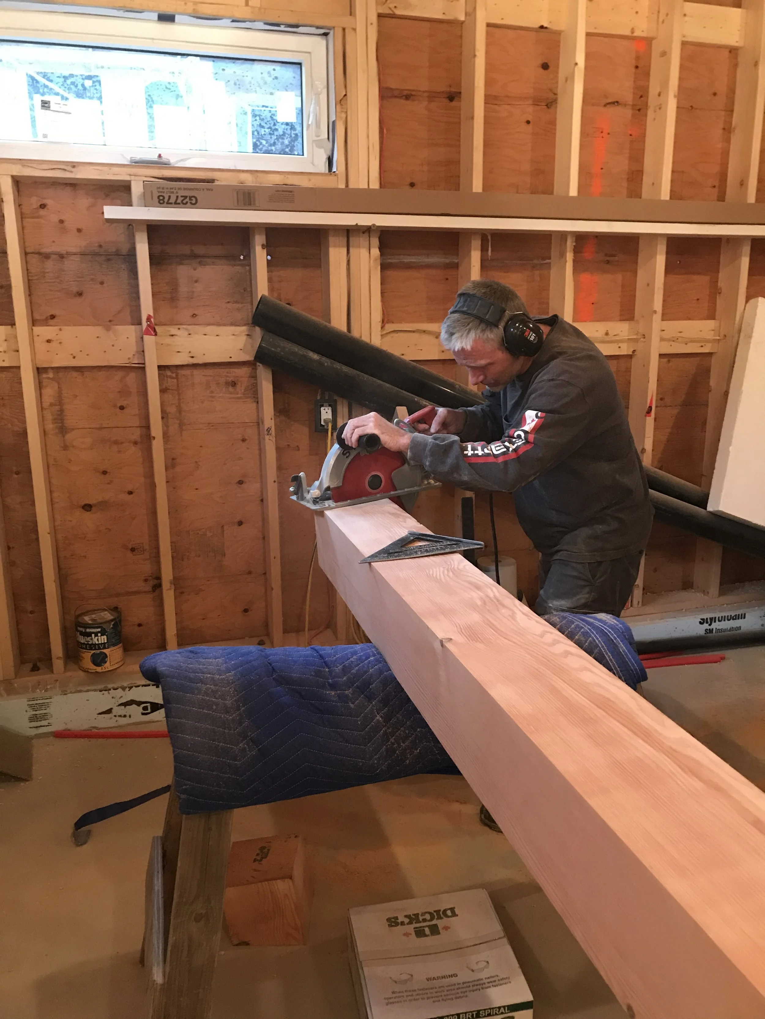 A man wearing ear protection and a dark hoodie is cutting a large piece of wood with a miter saw in a room under construction with unfinished wooden walls. There's a small window and a box of construction supplies on the floor.