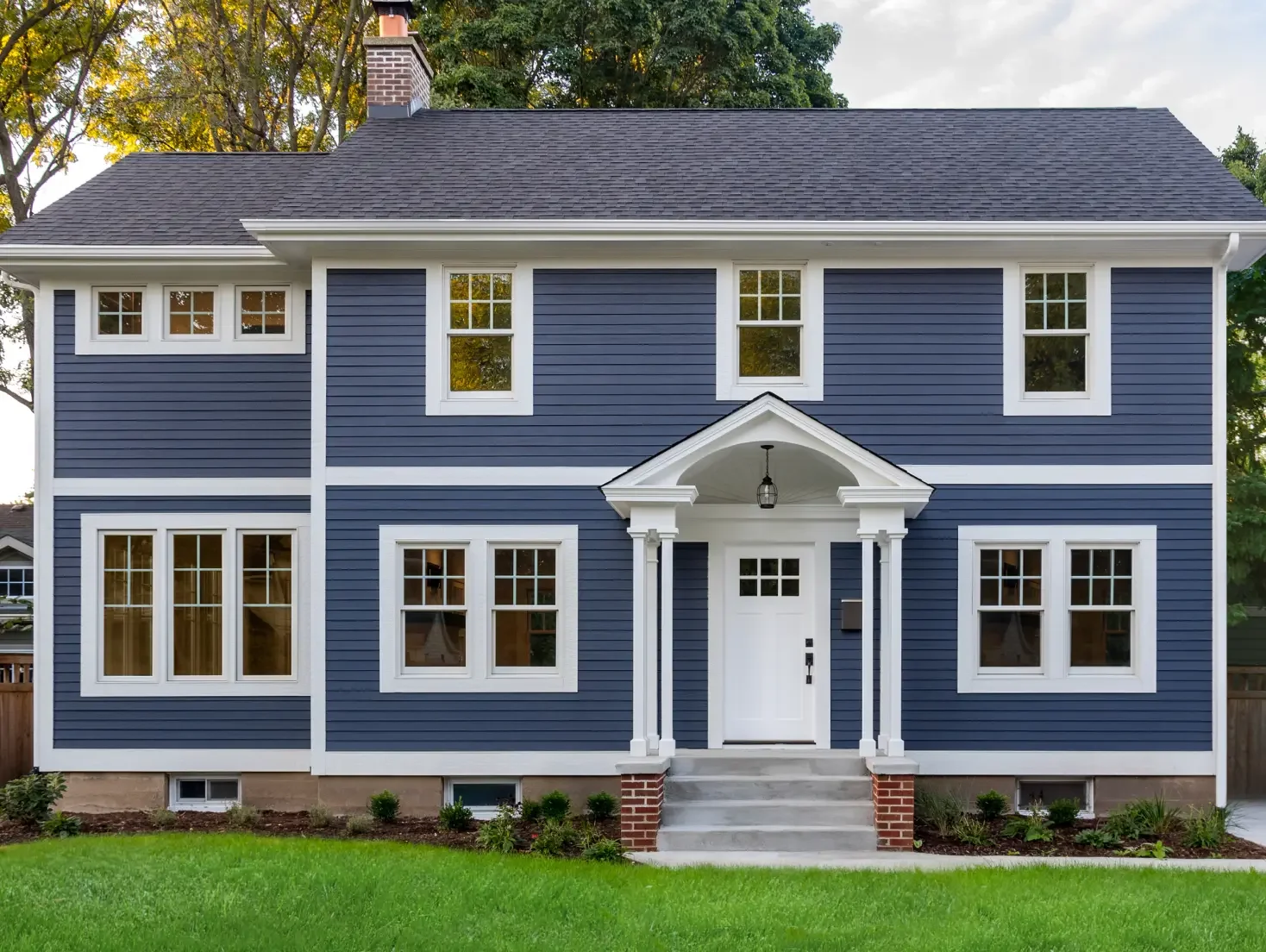A two-story house with blue siding, white trim, and a black roof, featuring multiple windows, a small front porch with steps, a mailbox, and a well-maintained lawn.