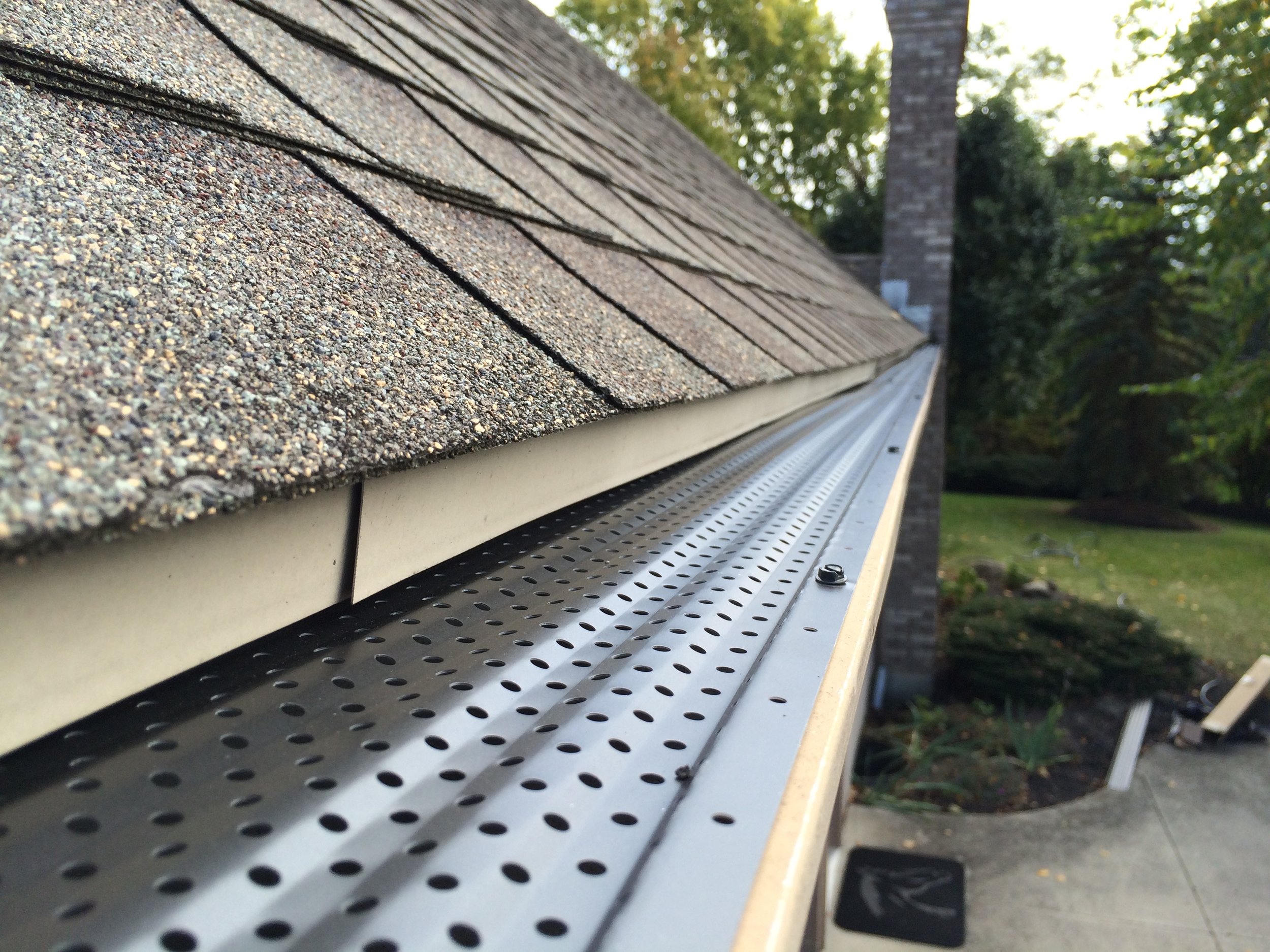 Close-up of a roof's gutter with perforated metal, shingles, and brick chimney in the background, surrounded by trees.