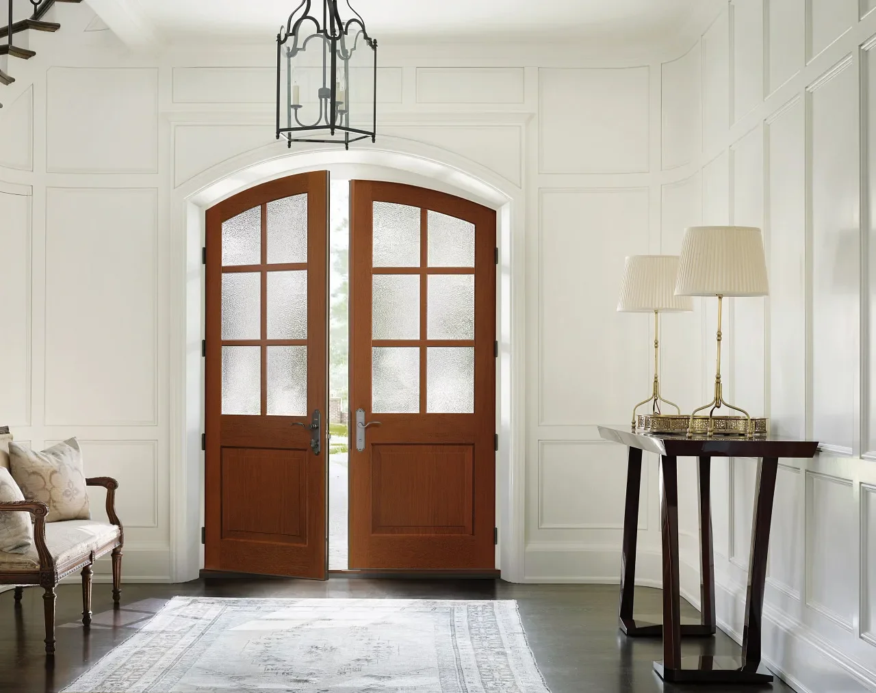 Interior entryway with double wooden doors and frosted glass panels, white paneled walls, a bench with cushions on the left, a small dark wood console table with two lamps on the right, a ceiling lantern light fixture, and a light-colored area rug on a dark hardwood floor.