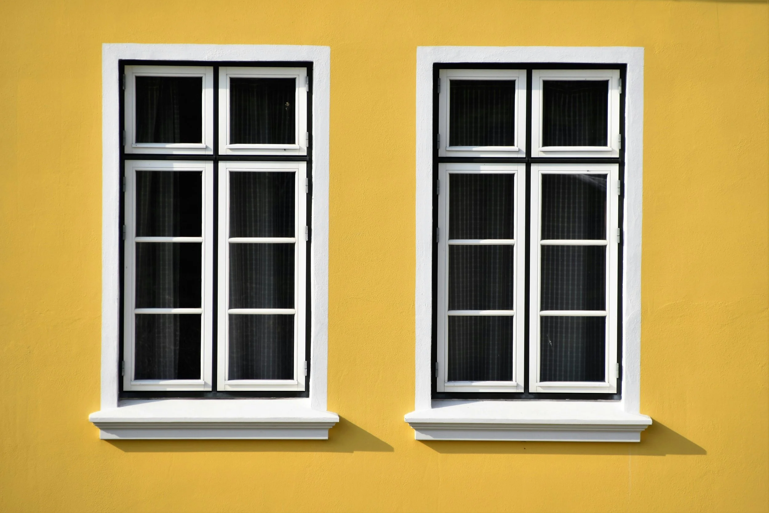 Two white-framed paneled windows on a yellow exterior wall of a building, with white window sills and black window panes.