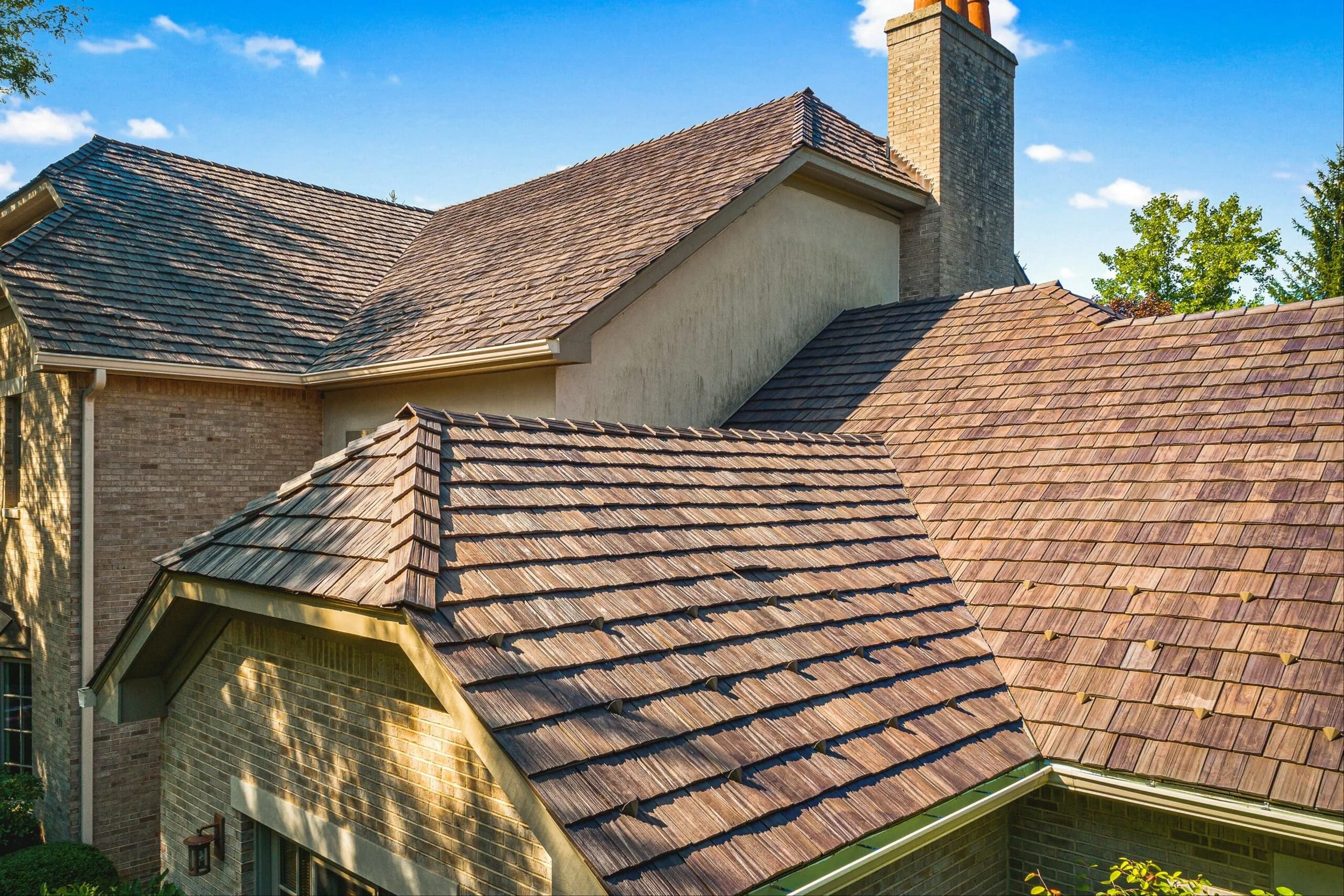 Multiple rooftops with shingles and chimneys on a house with brick and stucco exterior walls, under a blue sky with a few clouds.