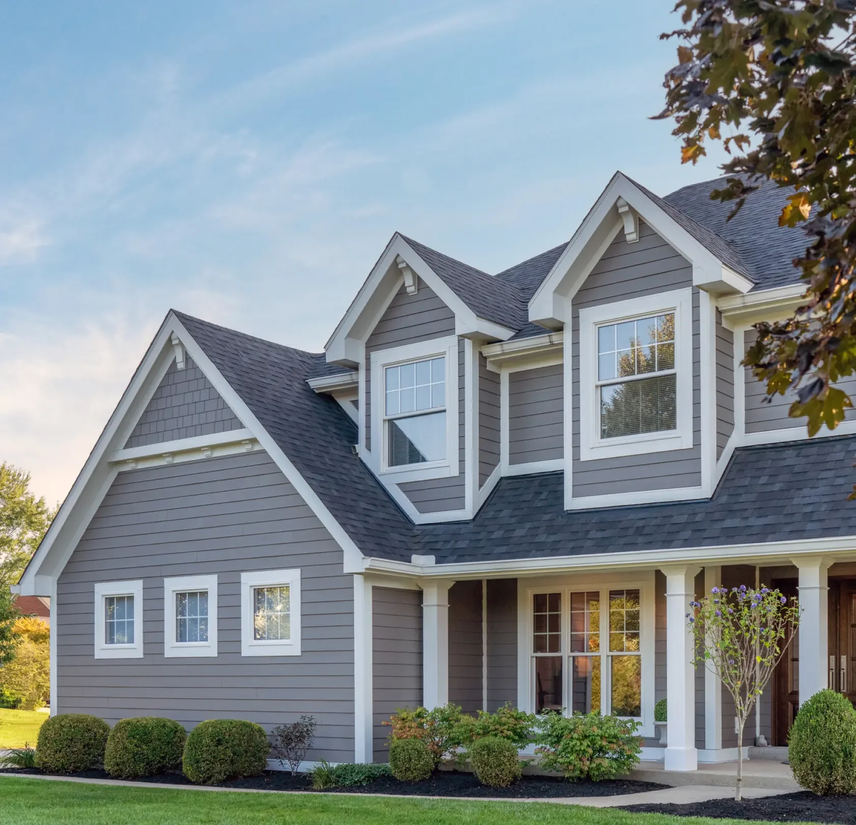 A modern gray house with white trim, multiple pitched roofs, and a front porch, surrounded by shrubs and a small tree, under a blue sky.