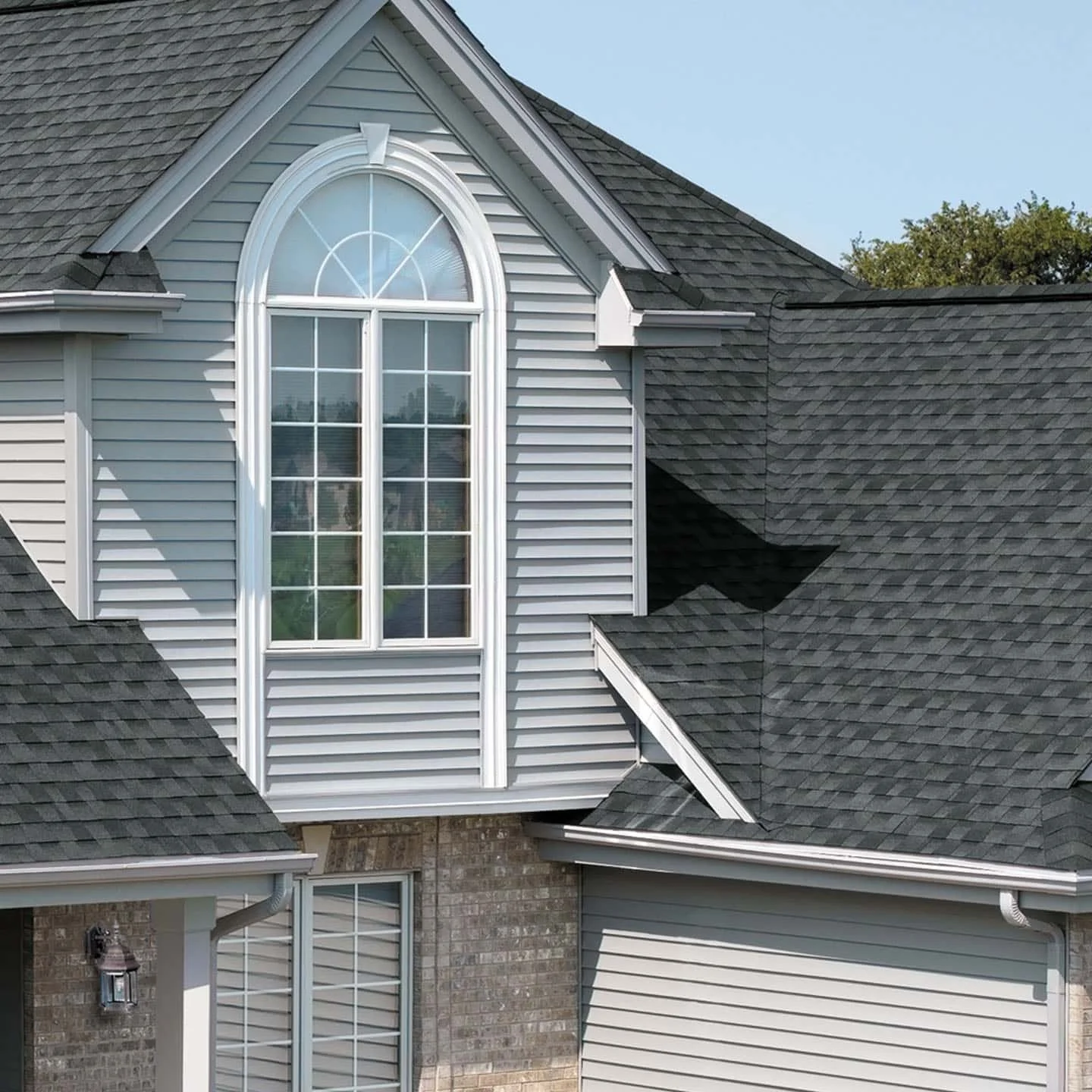 Close-up of a house's upper front section featuring a large arched window with multiple panes, grey vinyl siding, and a shingled roof.
