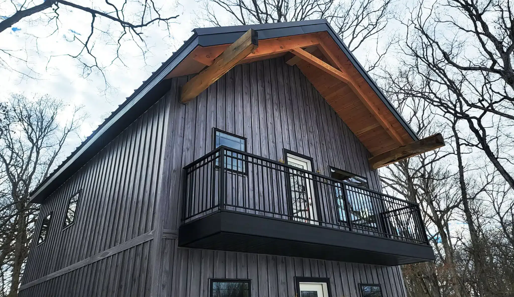 A three-story house with black wooden siding, large windows, and a black metal balcony on the second floor. The house has a steep gable roof with exposed wooden beams, and trees with bare branches surround it.