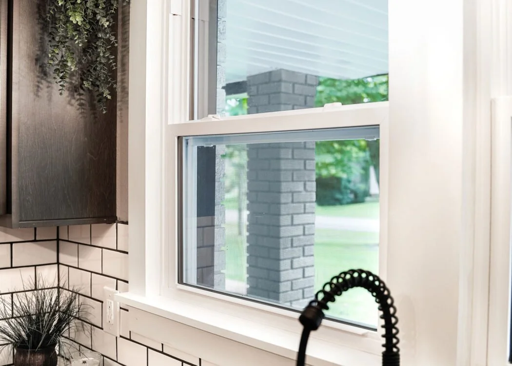 Close-up of a kitchen window with a view of an outdoor porch and greenery, including a brick pillar, white trim, and a black faucet in the foreground.