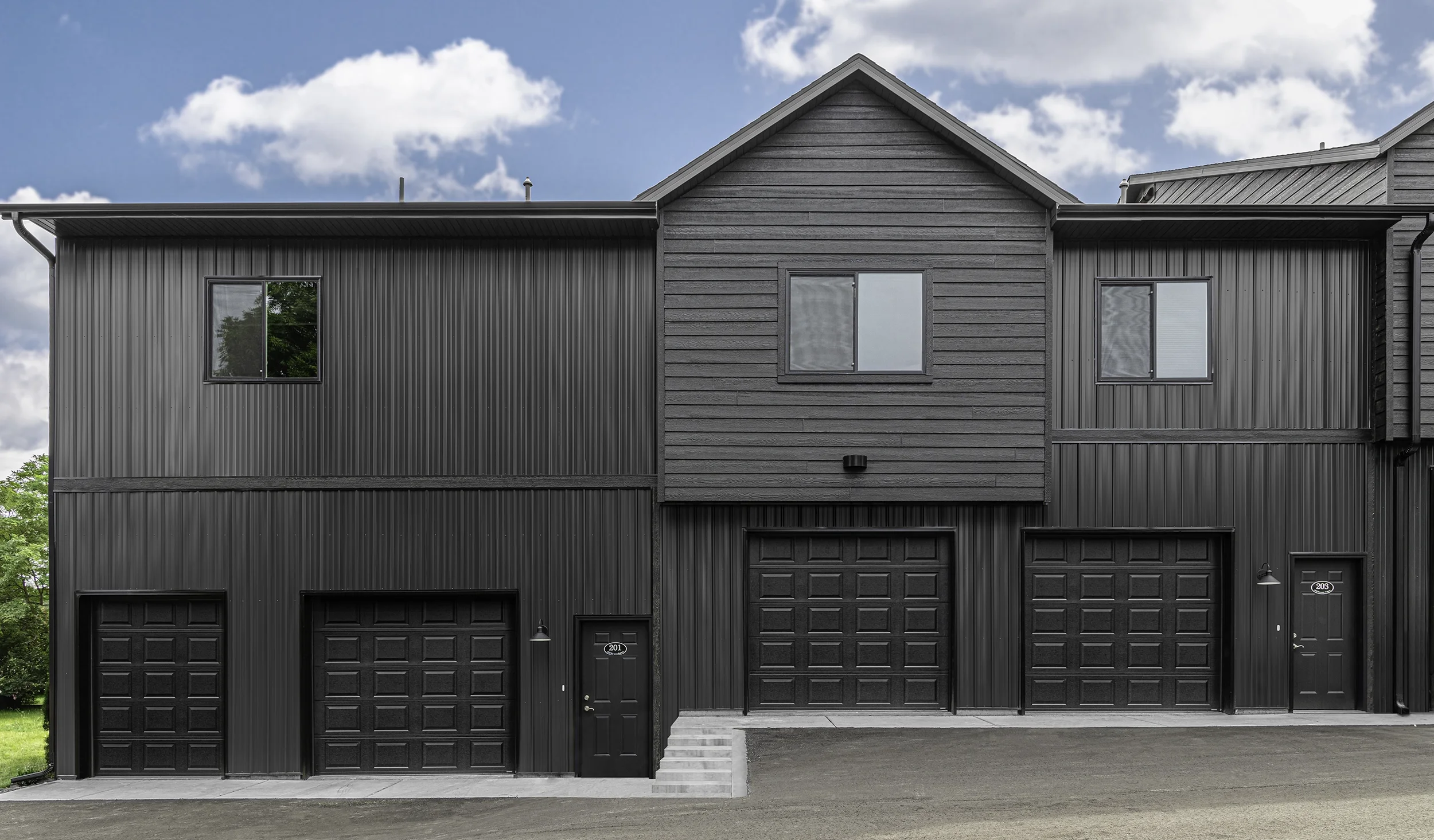 A modern black multi-family apartment building with three garage doors, two windows, and a door, with a blue sky and clouds above.