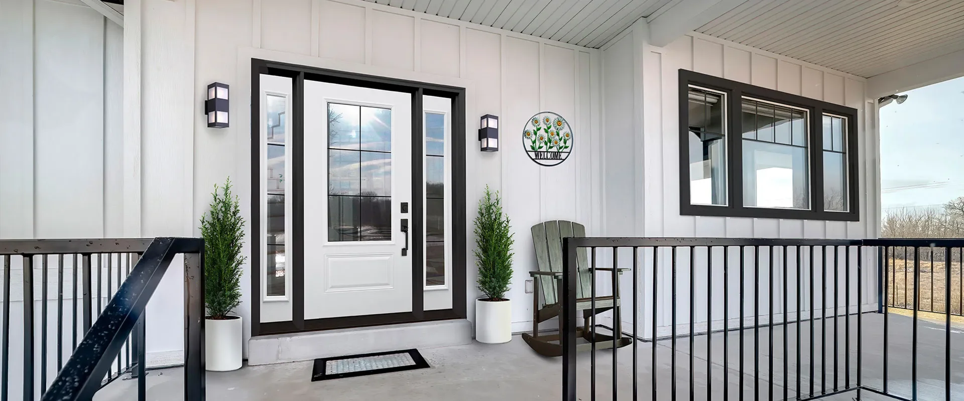 Front porch of a house with a white door, black trim, two potted plants, a wooden chair, a welcome sign, and large windows, with black railings and a concrete floor.