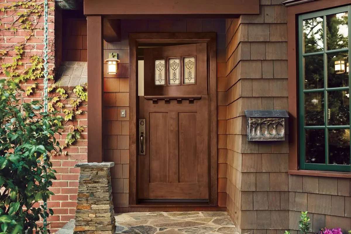 Front entrance of a house featuring a wooden door with decorative glass panels at the top, a stone walkway, brick and shingle siding, and a mailbox on the wall.
