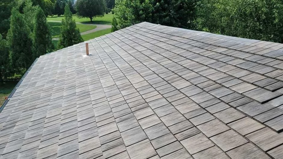 An aerial view of a house roof covered with gray shingles, surrounded by trees and a green landscape.
