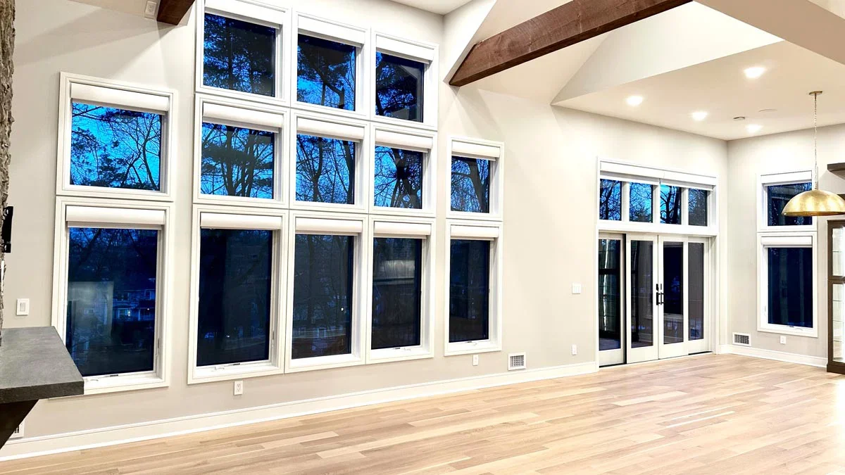 Empty modern living room with large windows and hardwood floors, showing trees outside at dusk, with ceiling lights and a hanging pendant light.