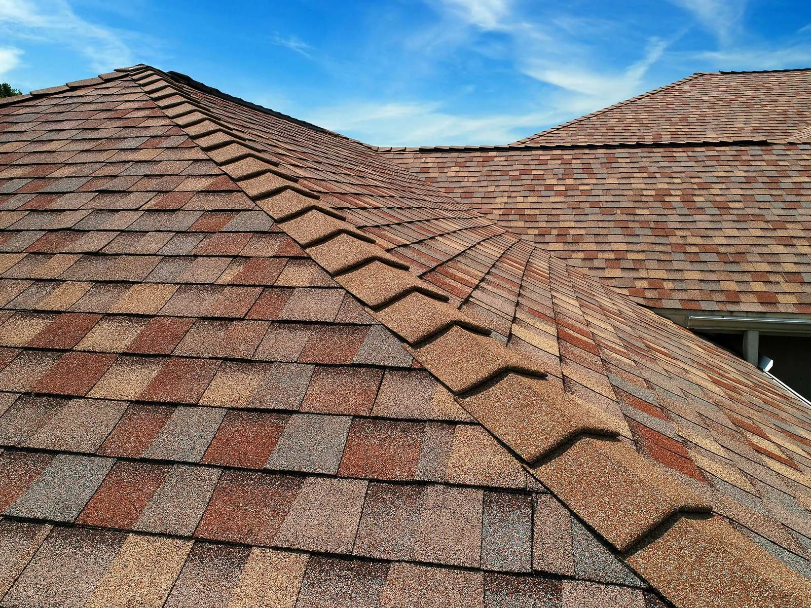 Close-up of a roof with reddish-brown asphalt shingles under a blue sky with light clouds.
