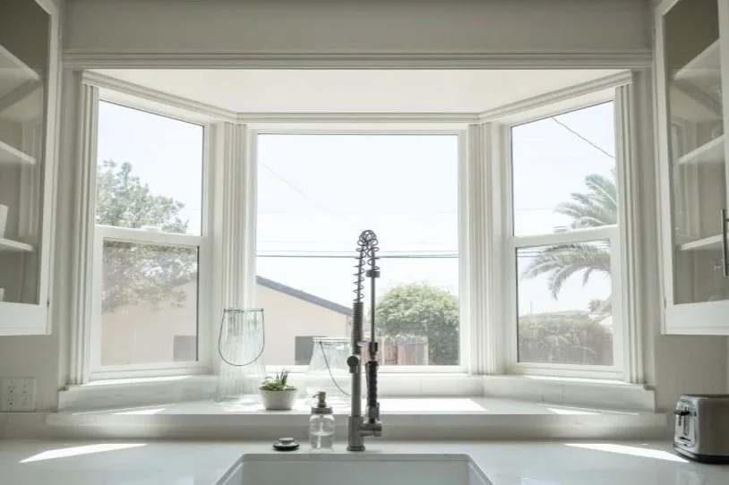 Kitchen sink with faucet, soap dispenser, small plant, and glass jar by a large window with garden and palm tree outside