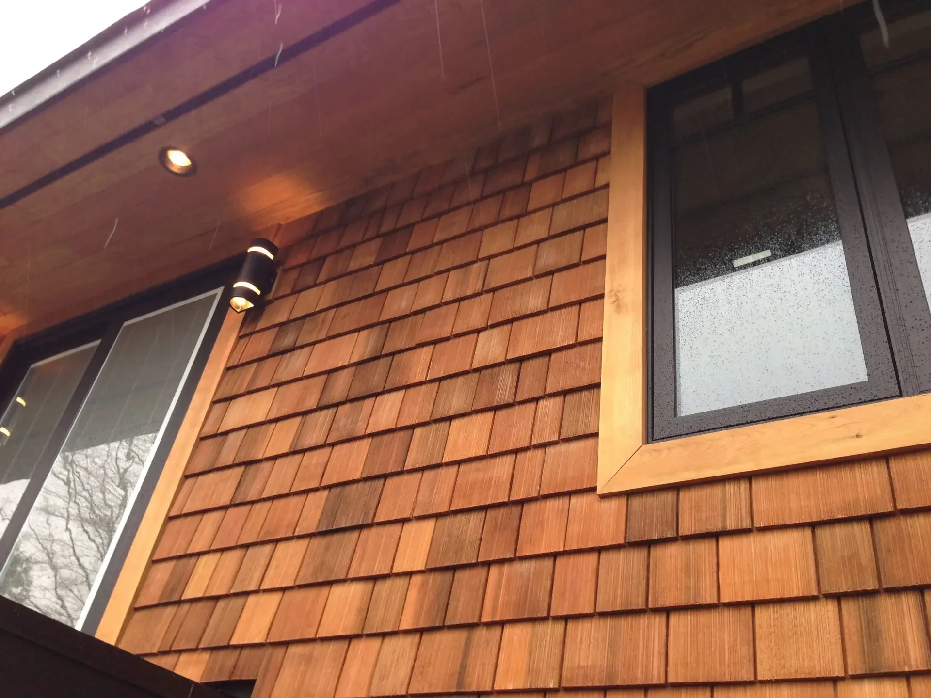 A wooden house exterior with a window and outdoor lighting, captured during rainy weather.