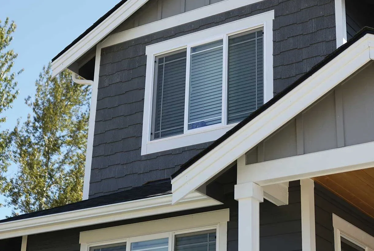 Close-up of a modern two-story house with dark gray siding, white trim, and double-pane window with blinds, with trees and clear blue sky in the background.