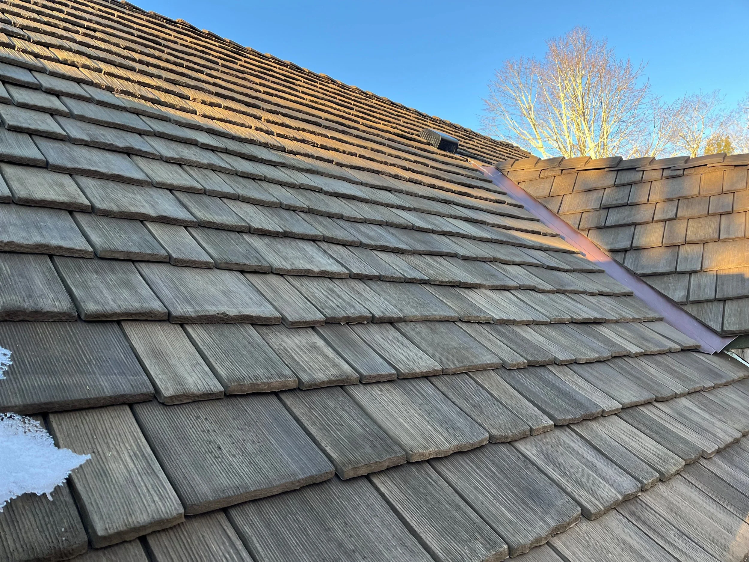 Close-up view of a wooden shingled roof with different sections showing weathered and new shingles, under a clear blue sky with bare trees in the background.